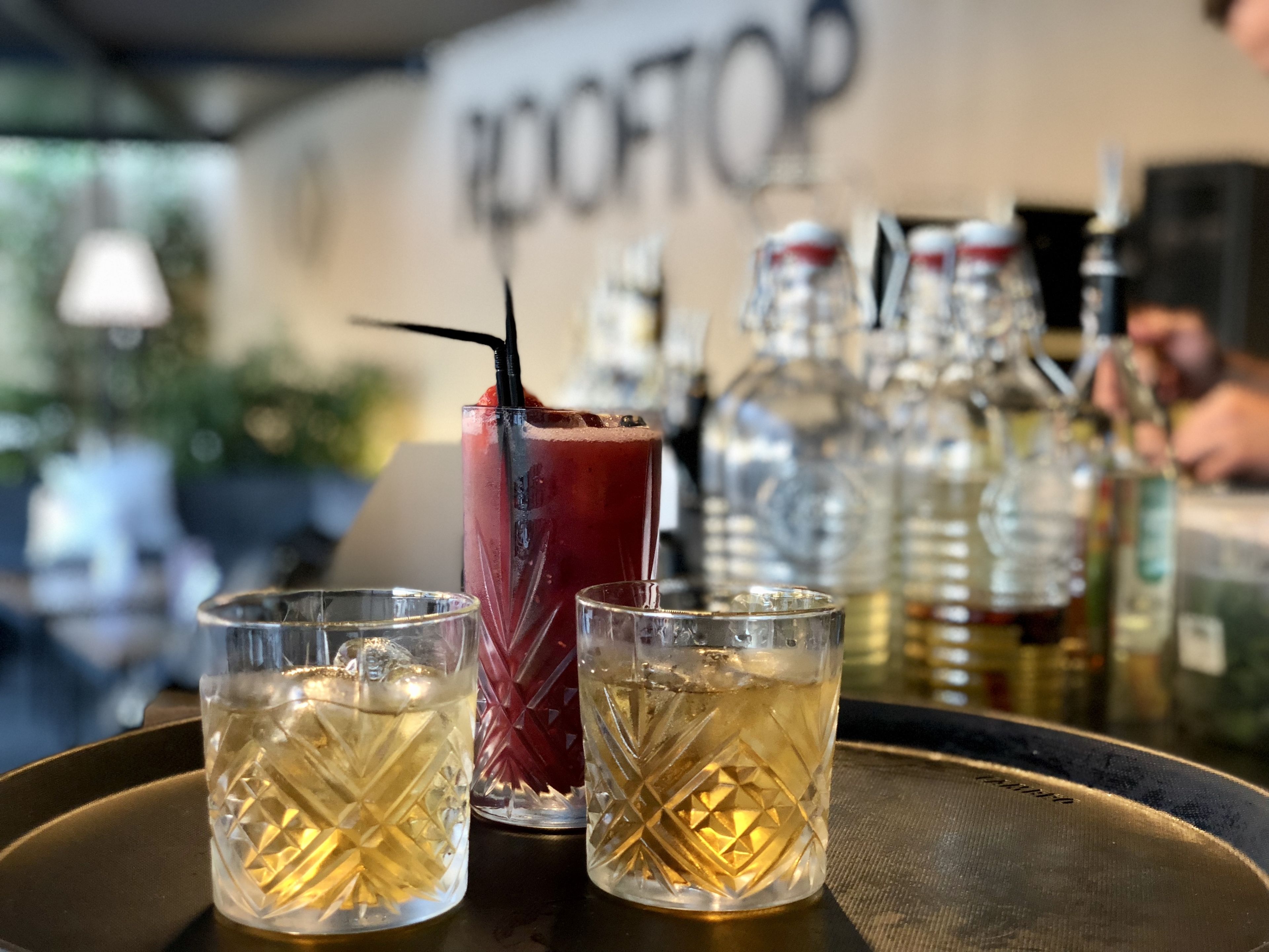 A bar area with three glasses of a golden beverage, one with a red drink and a black straw, and several bottles in the background.