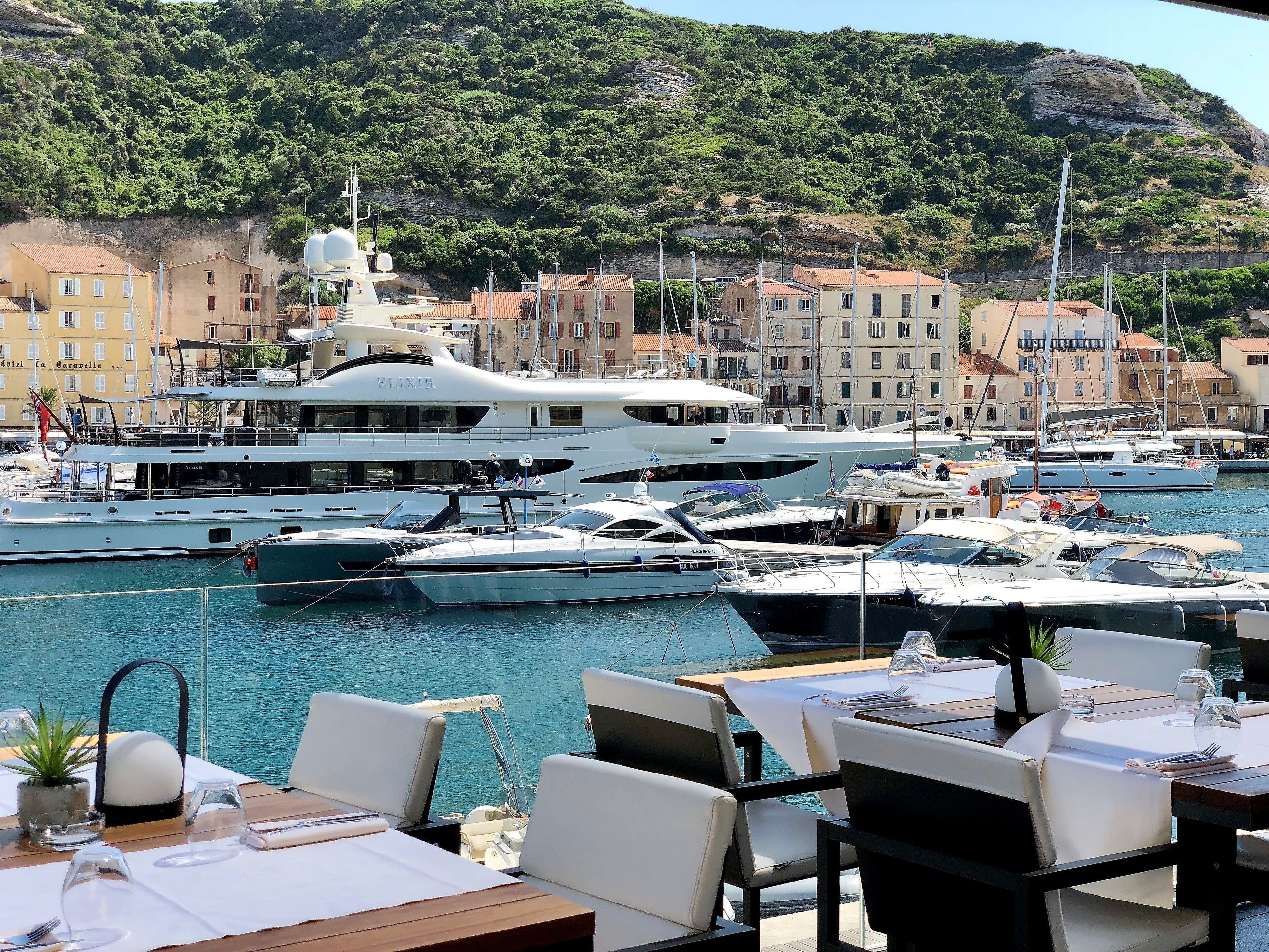 Een jachthaven met aangemeerde zeilboten, een restaurant met terras en gebouwen op de achtergrond.