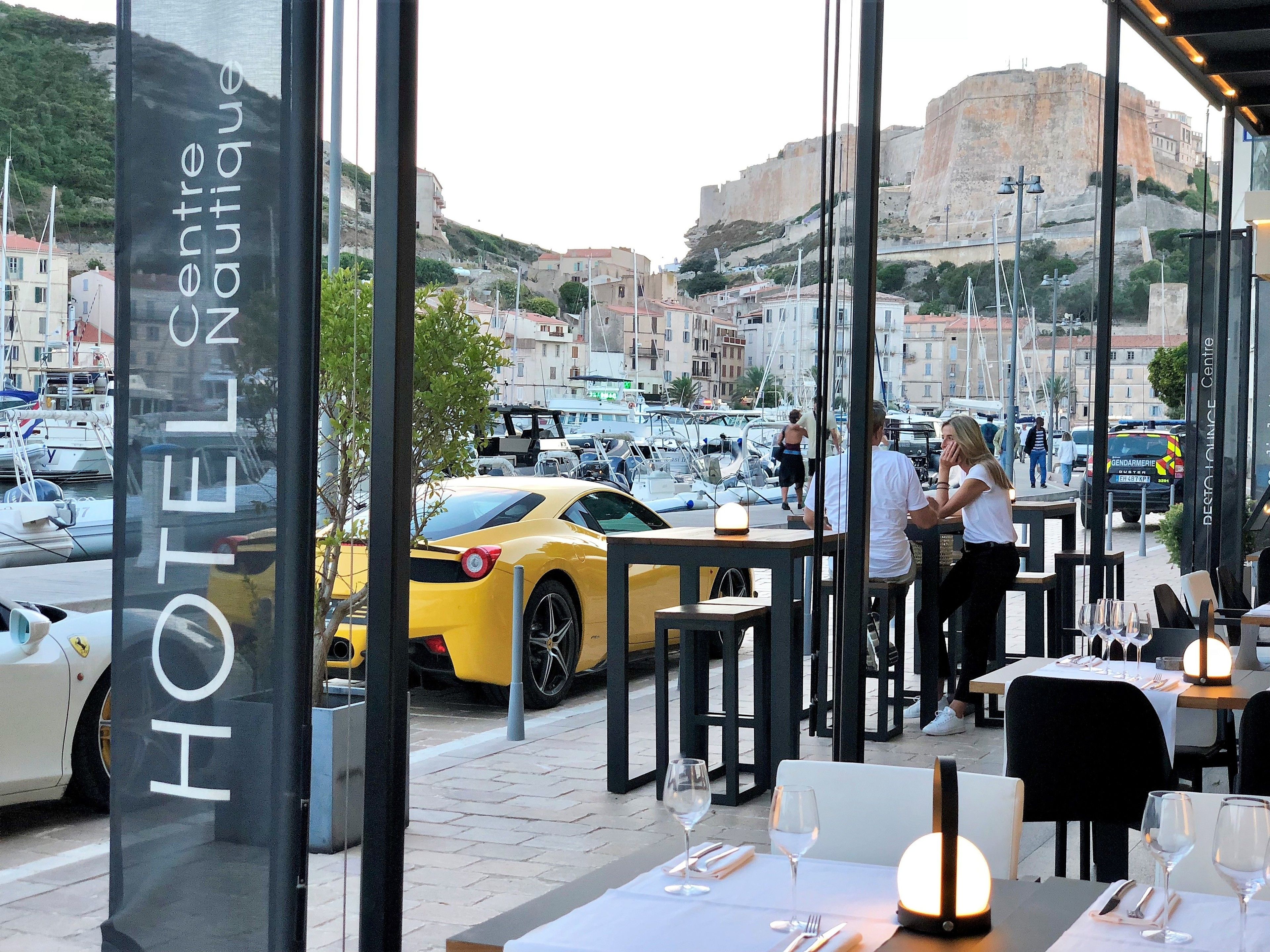 A restaurant with outdoor seating overlooking a marina, featuring a yellow sports car parked nearby.