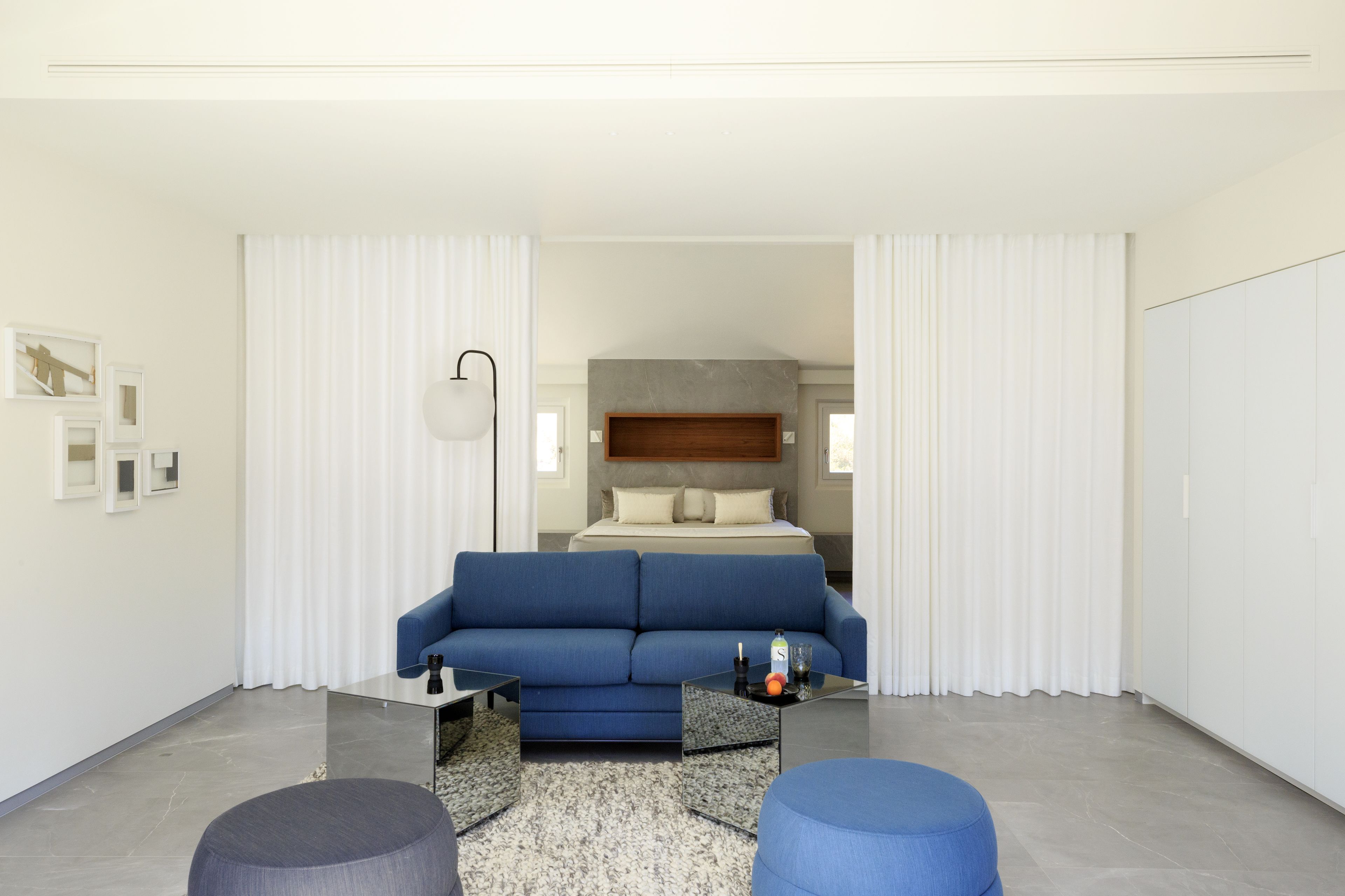 Ein modernes Hotelzimmer mit einem blauen Sofa, einem Glastisch und einem runden Ottomanen. Ein Bett mit Holz Kopfteil und weißen Vorhängen ist ebenfalls vorhanden.