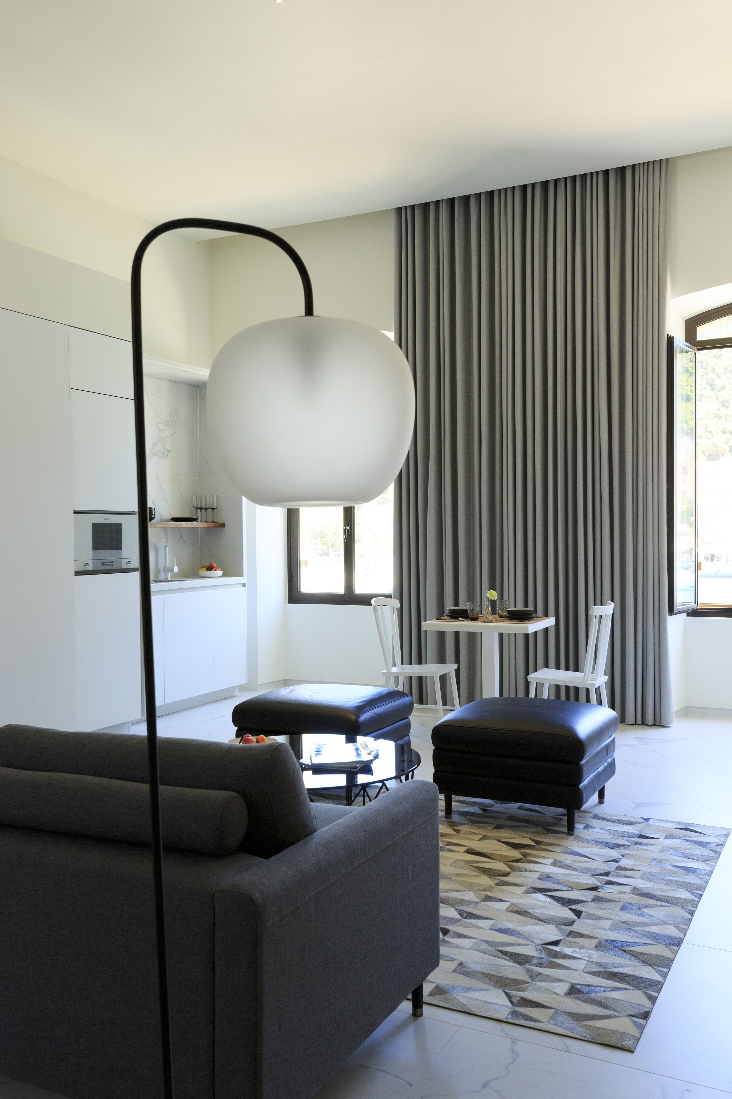A modern living room with a grey sofa, a black ottoman, a dining table with chairs, and a large window with curtains.