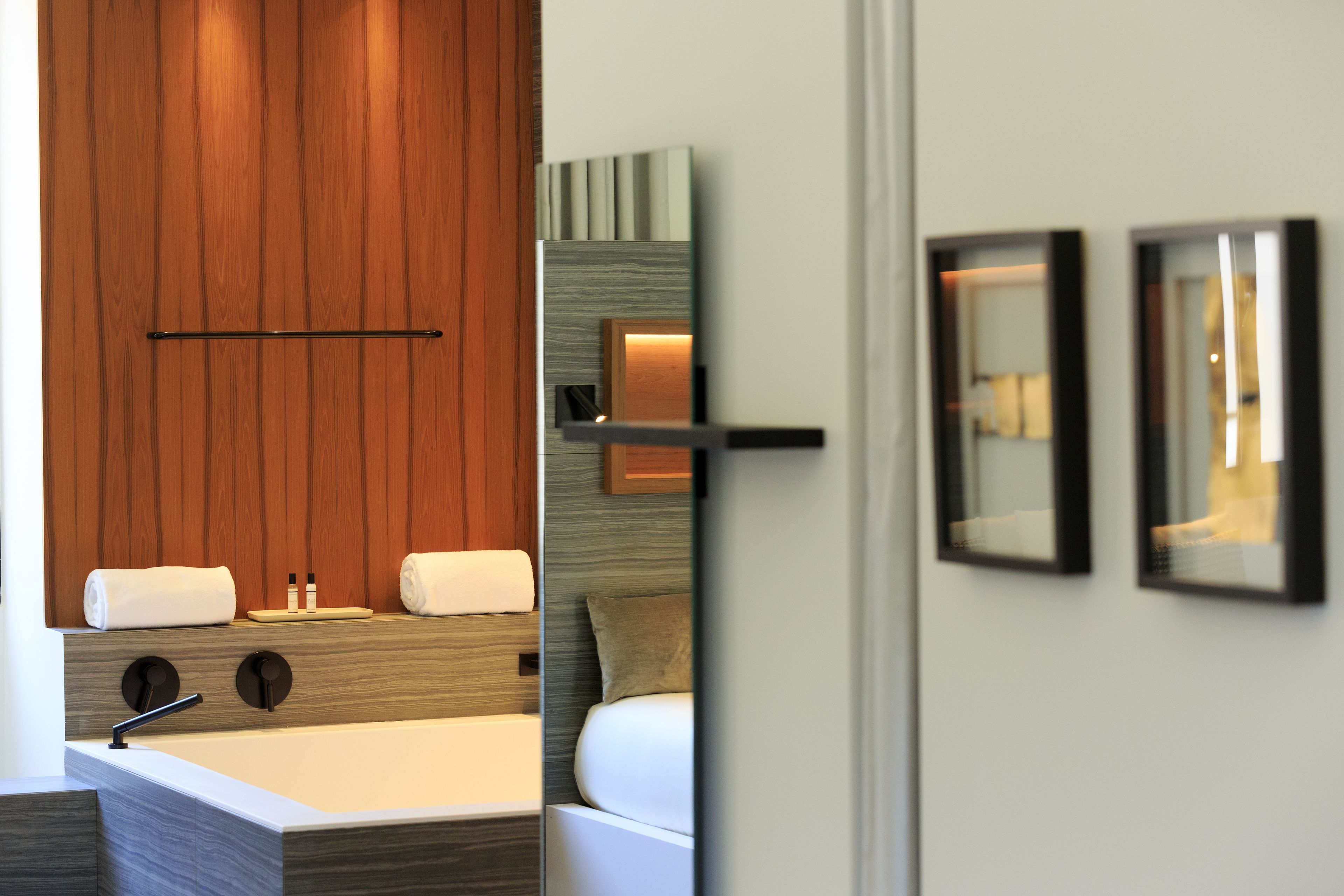 A modern hotel room with a wooden panel wall, a large mirror, a bed with white bedding, and a small bedside table.