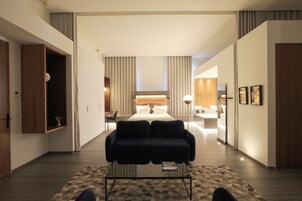 Una habitación de hotel moderna con una cama grande, un sofá oscuro, una mesa de centro de vidrio y vistas al pasillo.