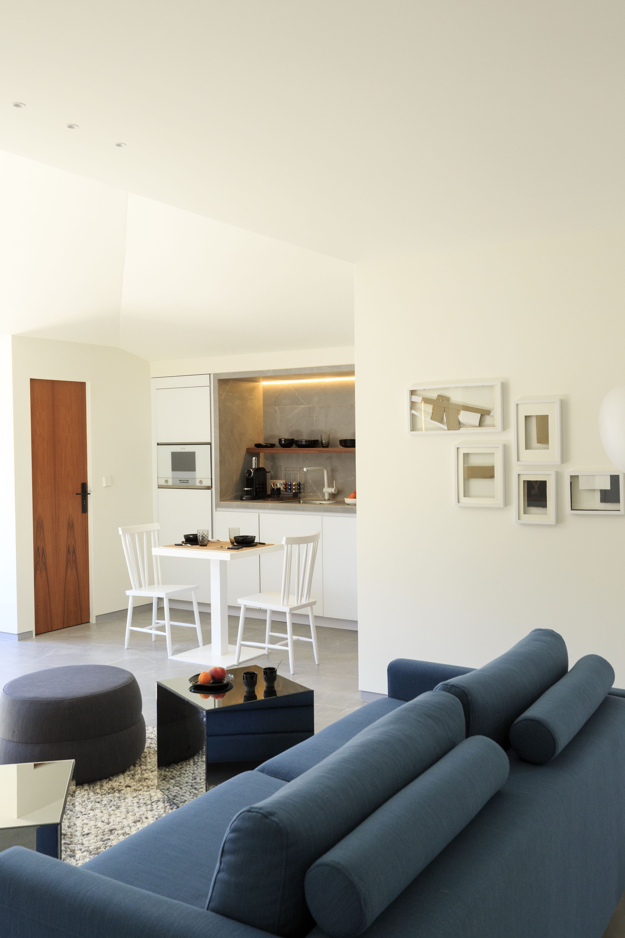 A modern living room with a blue sofa, a dining table with chairs, and a kitchen area visible through an open door.