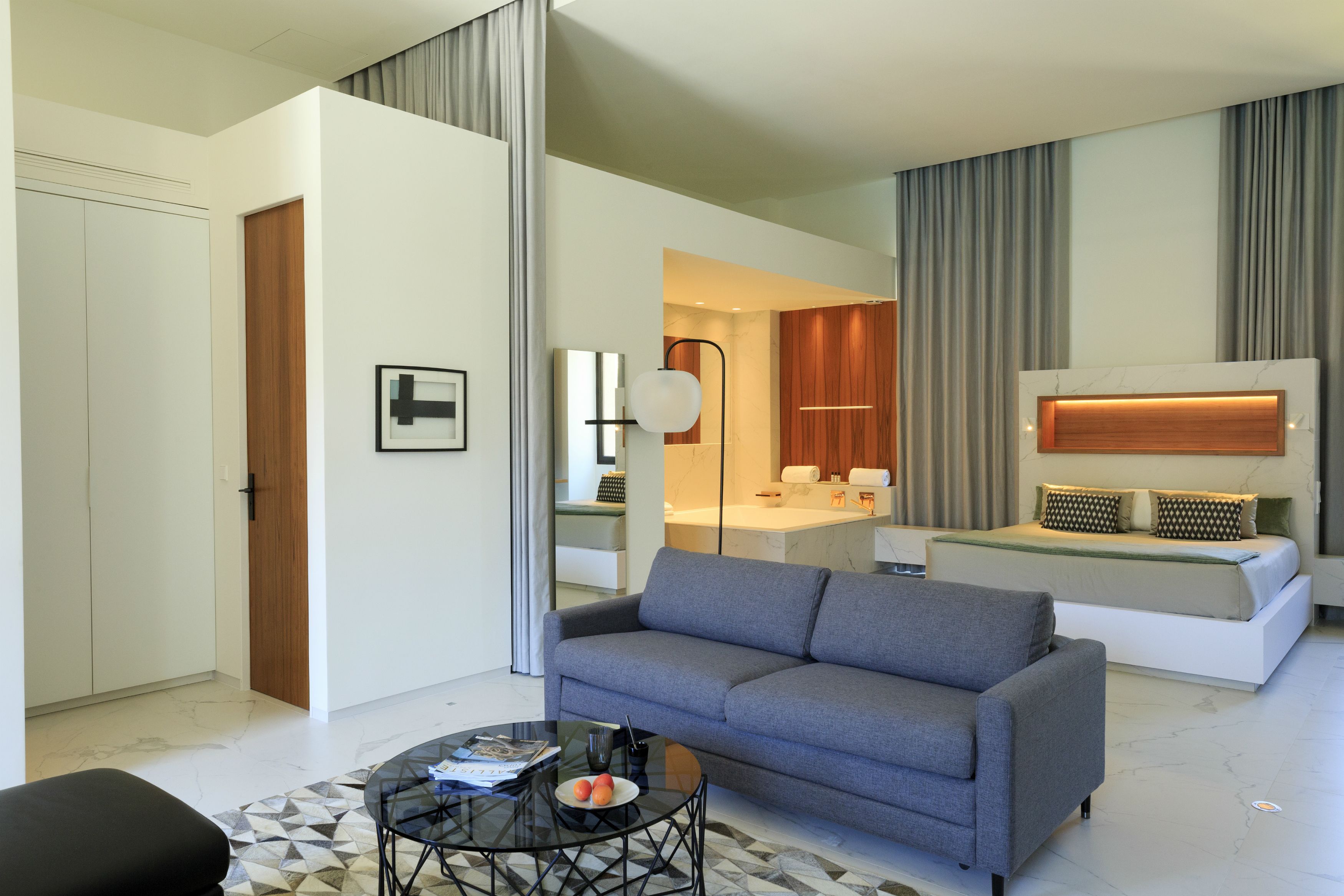 A modern living room with a grey sofa, a glass coffee table, and a black ottoman. There is a kitchen area with wooden cabinets and a white countertop visible through an open door.