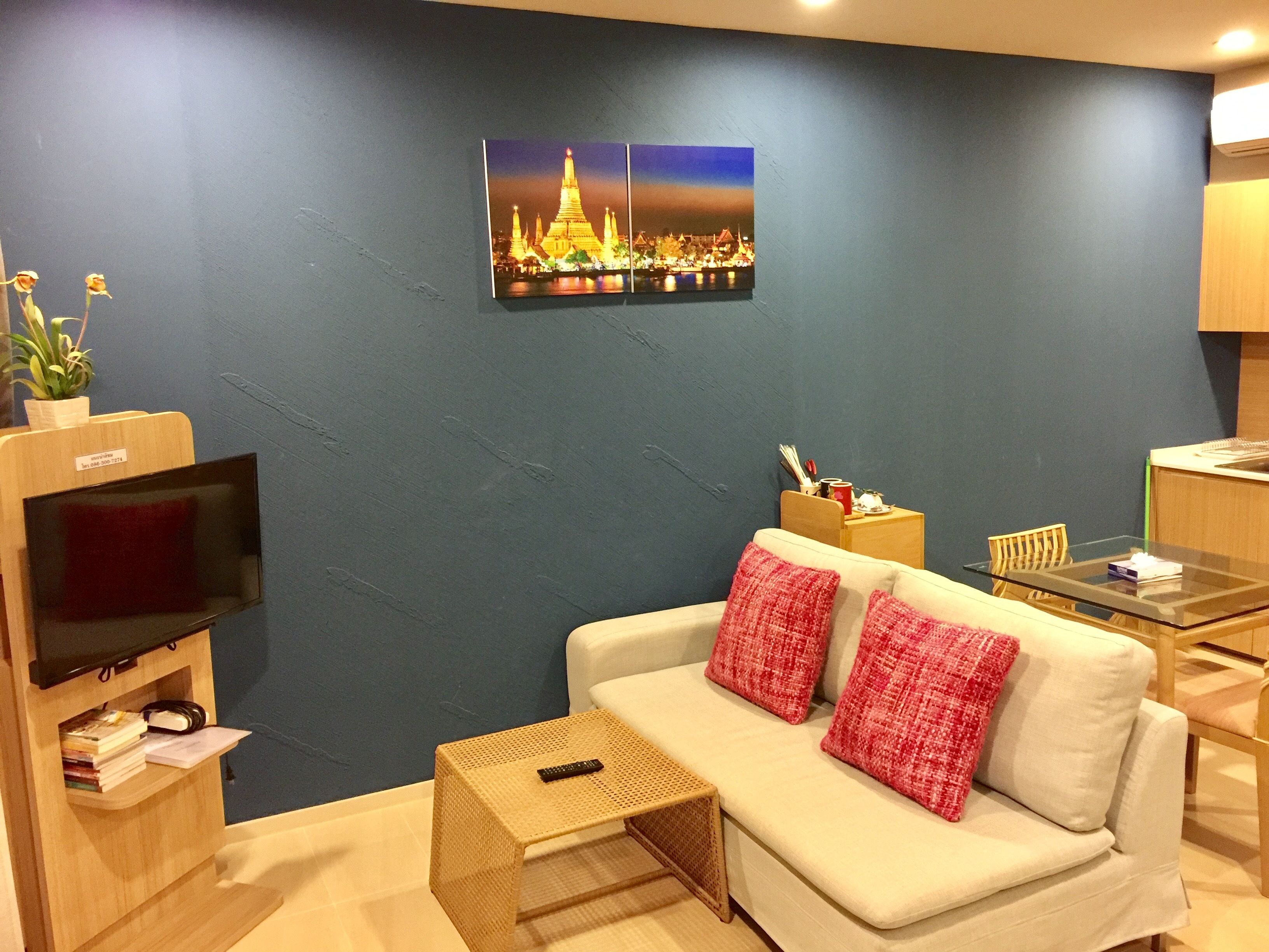A modern living room with a blue accent wall, a flat-screen TV, a white sofa with red cushions, a small coffee table, and a framed picture of a temple at night.