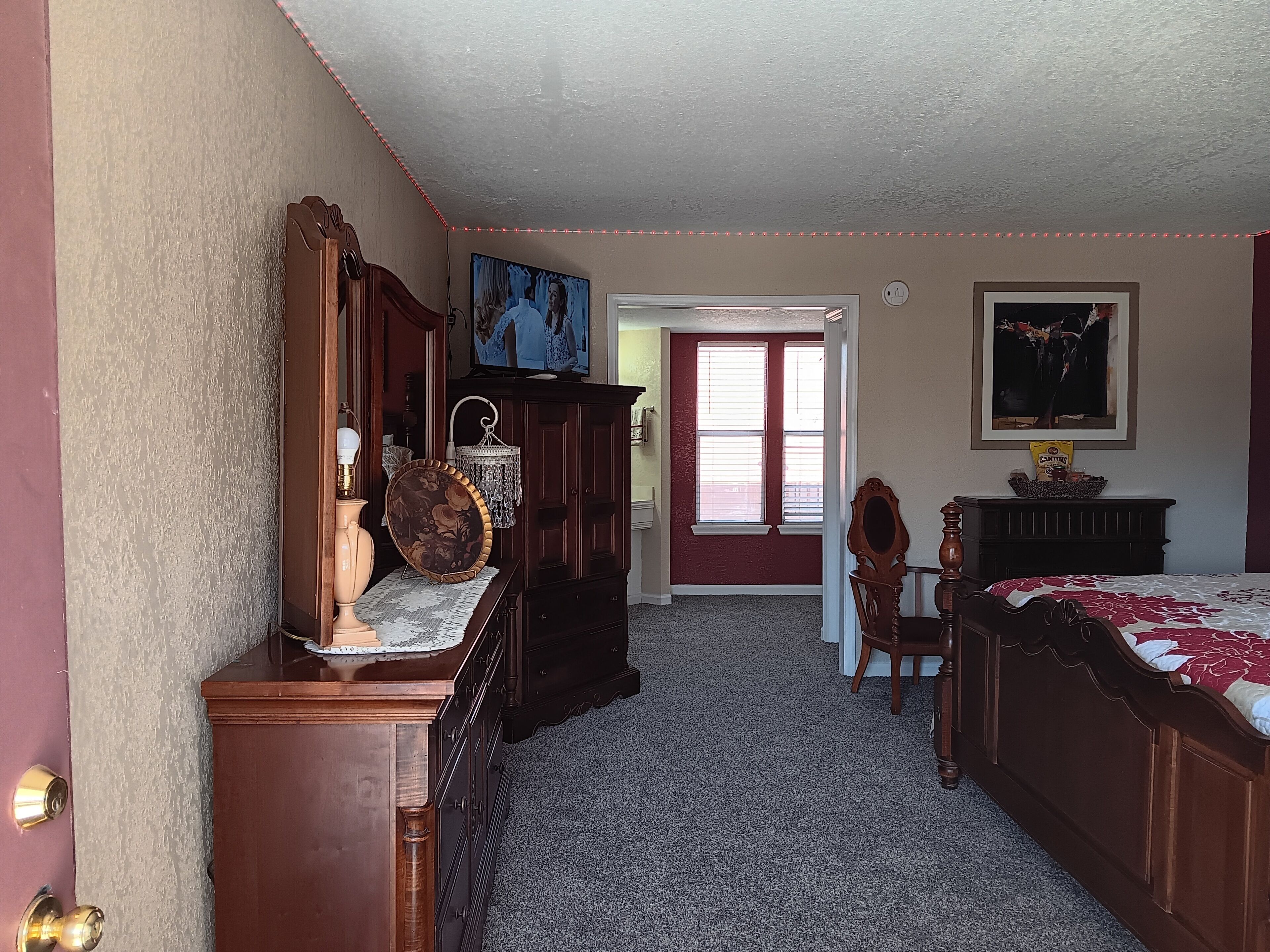 Une chambre avec une commode en bois, un lit recouvert d’une couvre-lit à motifs floraux, un lustre et un tableau accroché au mur.