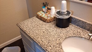 A bathroom with a granite countertop, a white sink, a mirror, a trash can, and a coffee maker.