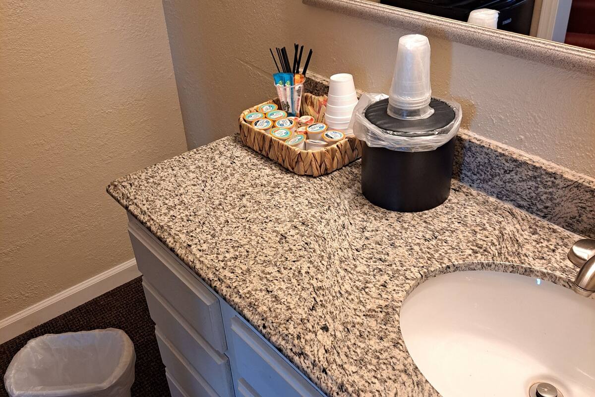 A bathroom with a granite countertop, a white sink, a mirror, a trash can, and a coffee maker.