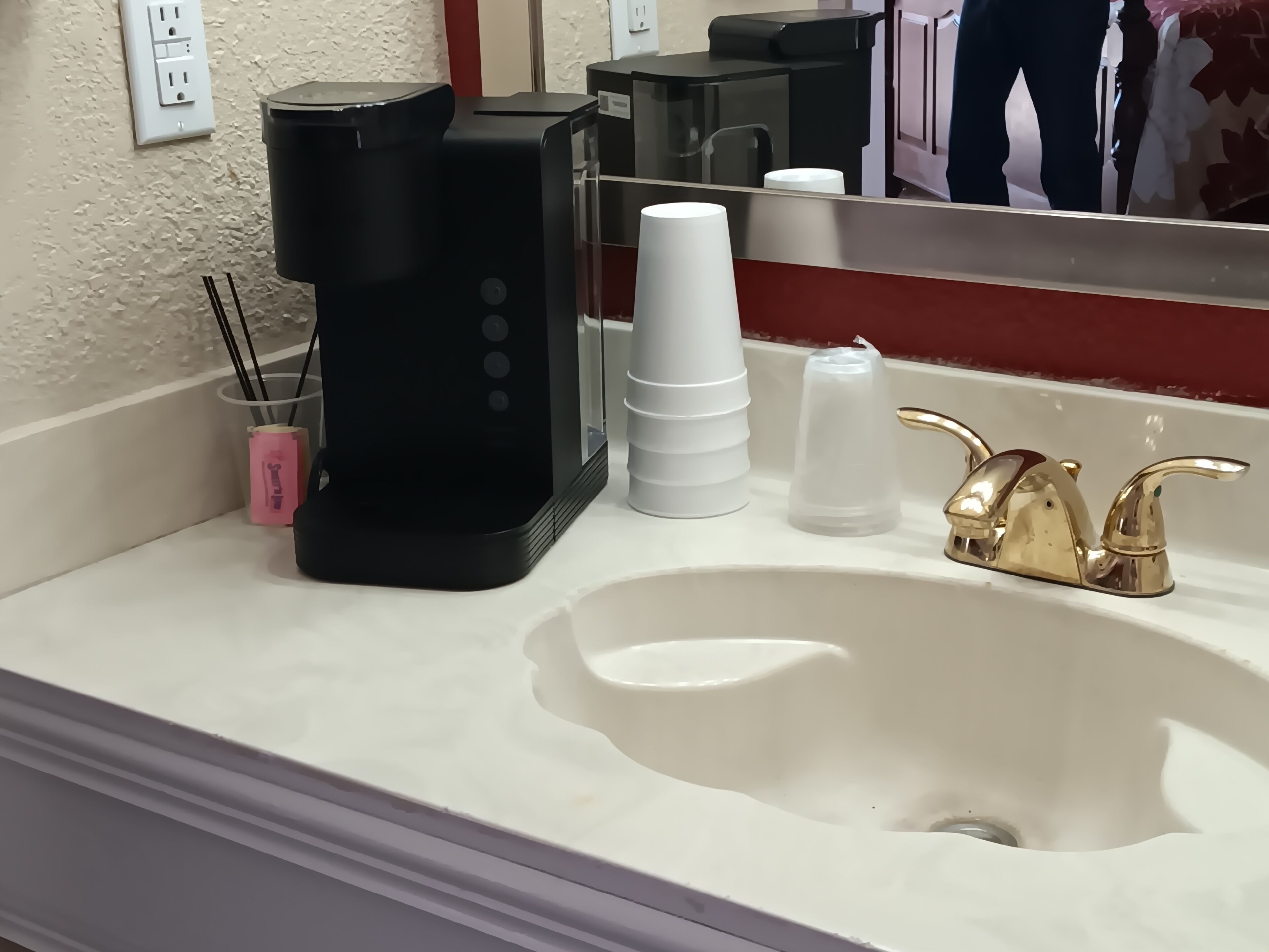 Een badkamer met een witte wastafel, een zwarte koffiezetapparaat, een spiegel en een gouden kraan.