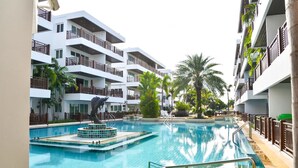 A swimming pool surrounded by multi-story buildings with balconies, palm trees, and a clear sky.