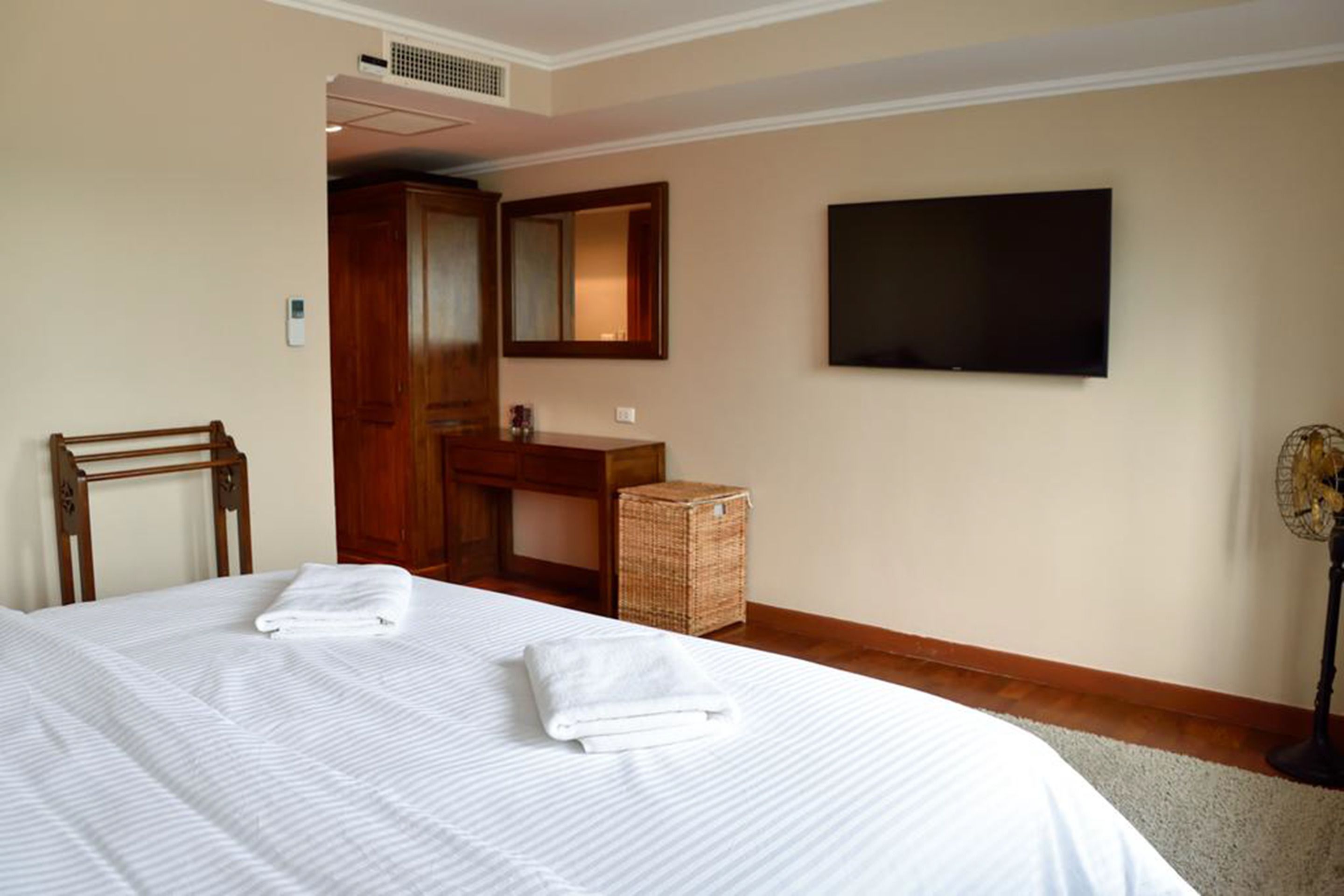 Une chambre d’hôtel avec un lit, une chaise en bois, une petite table, un miroir, une télévision et un ventilateur.