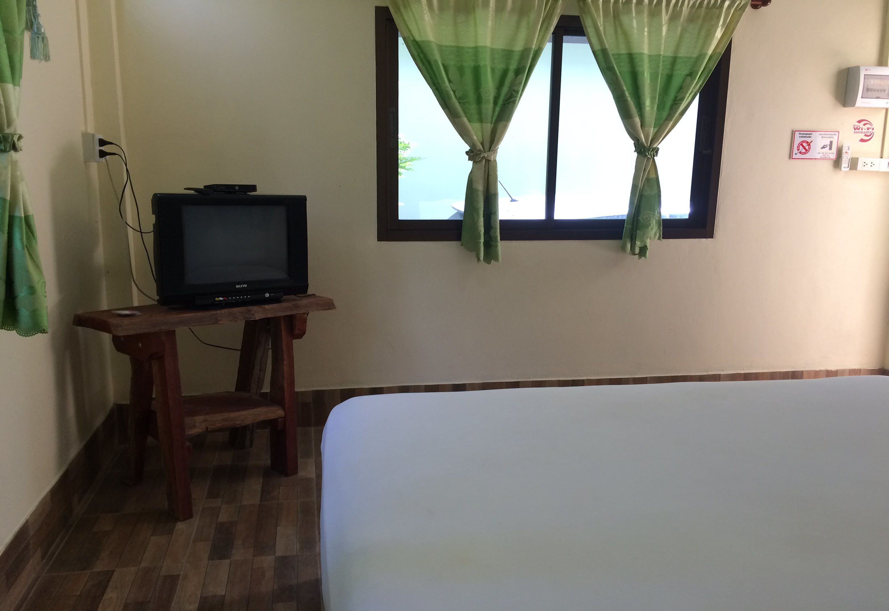 A room with a bed, a television on a wooden stand, a window with curtains, and a wall-mounted thermostat.