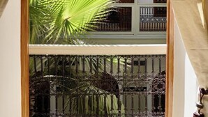 A view through a window with a palm tree, wooden balcony, and wrought iron railing. - Riad Tzarra (Marrakech)