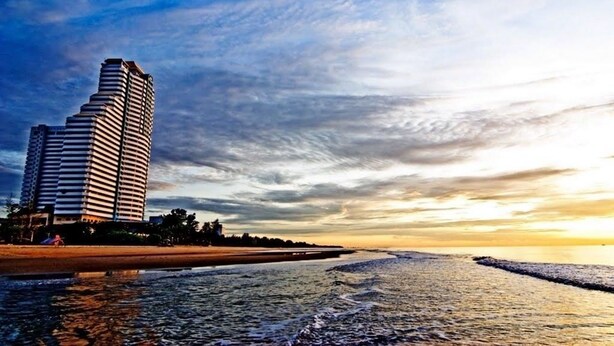 A high-rise hotel with a curved facade, situated by a beach with a calm sea and a sunset sky. - Cha-Am Grand Condotel (Cha-am)