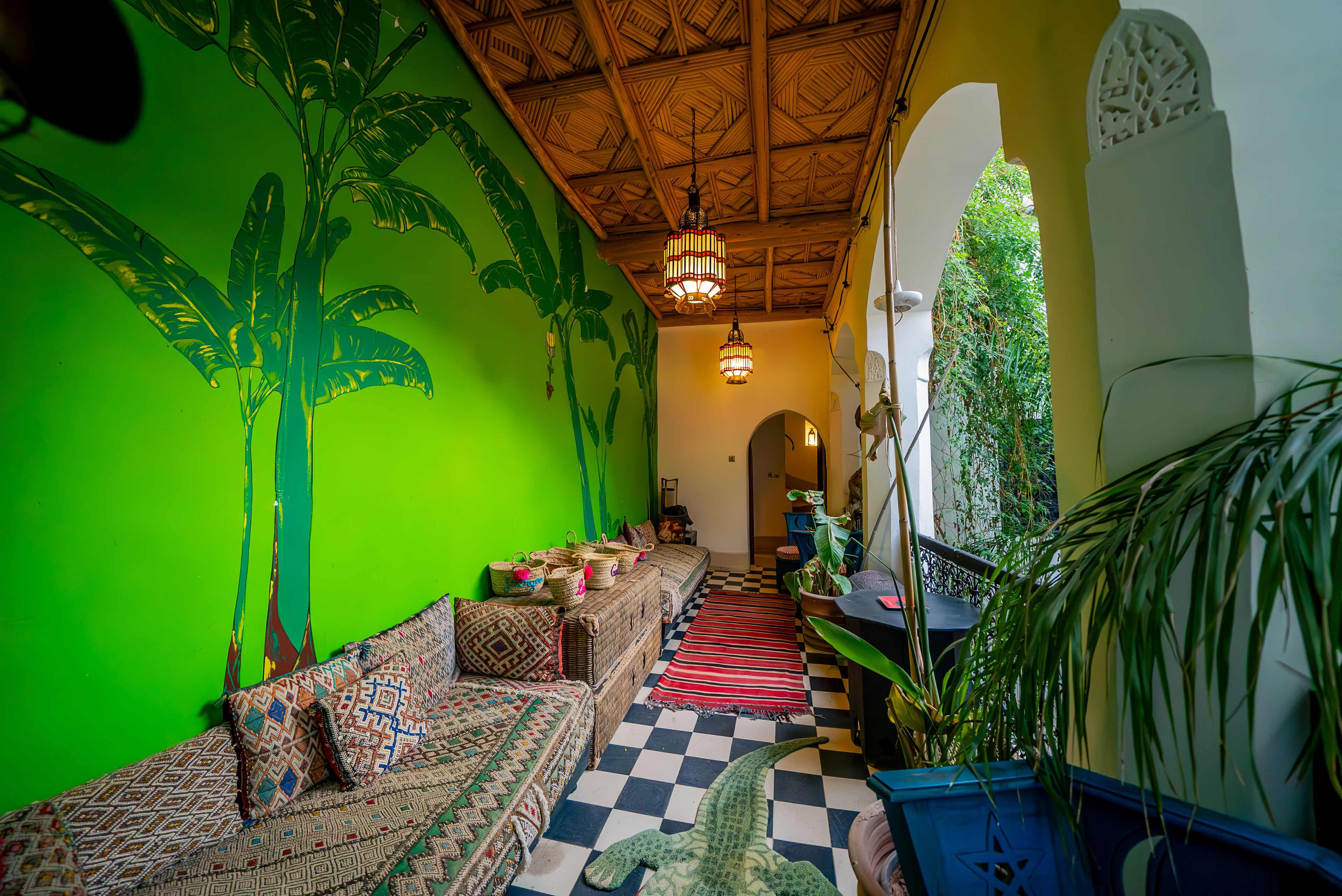 A room with a checkered floor, a green wall with a banana tree mural, a woven sofa, and a hanging light fixture.