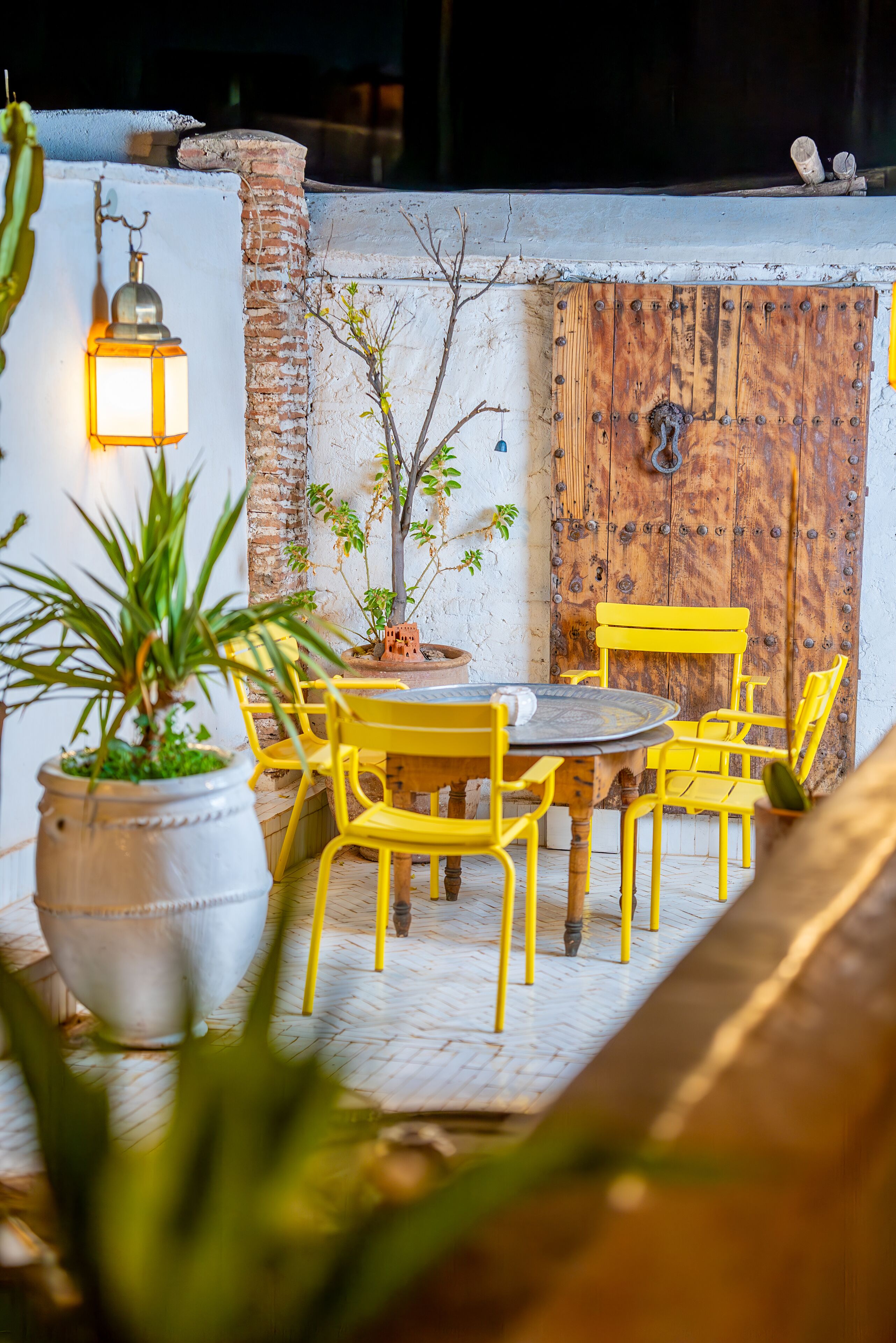 An outdoor seating area with yellow chairs and a table, surrounded by potted plants and a brick wall.