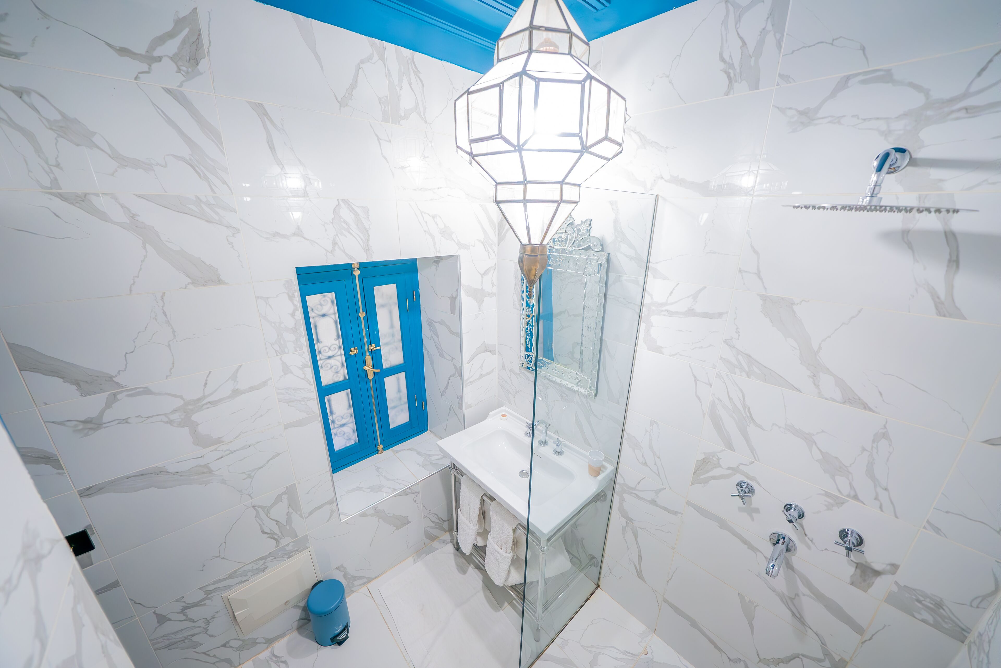 Un baño con paredes de mármol blanco, una puerta azul, ducha con mampara de vidrio y lavamanos blanco.