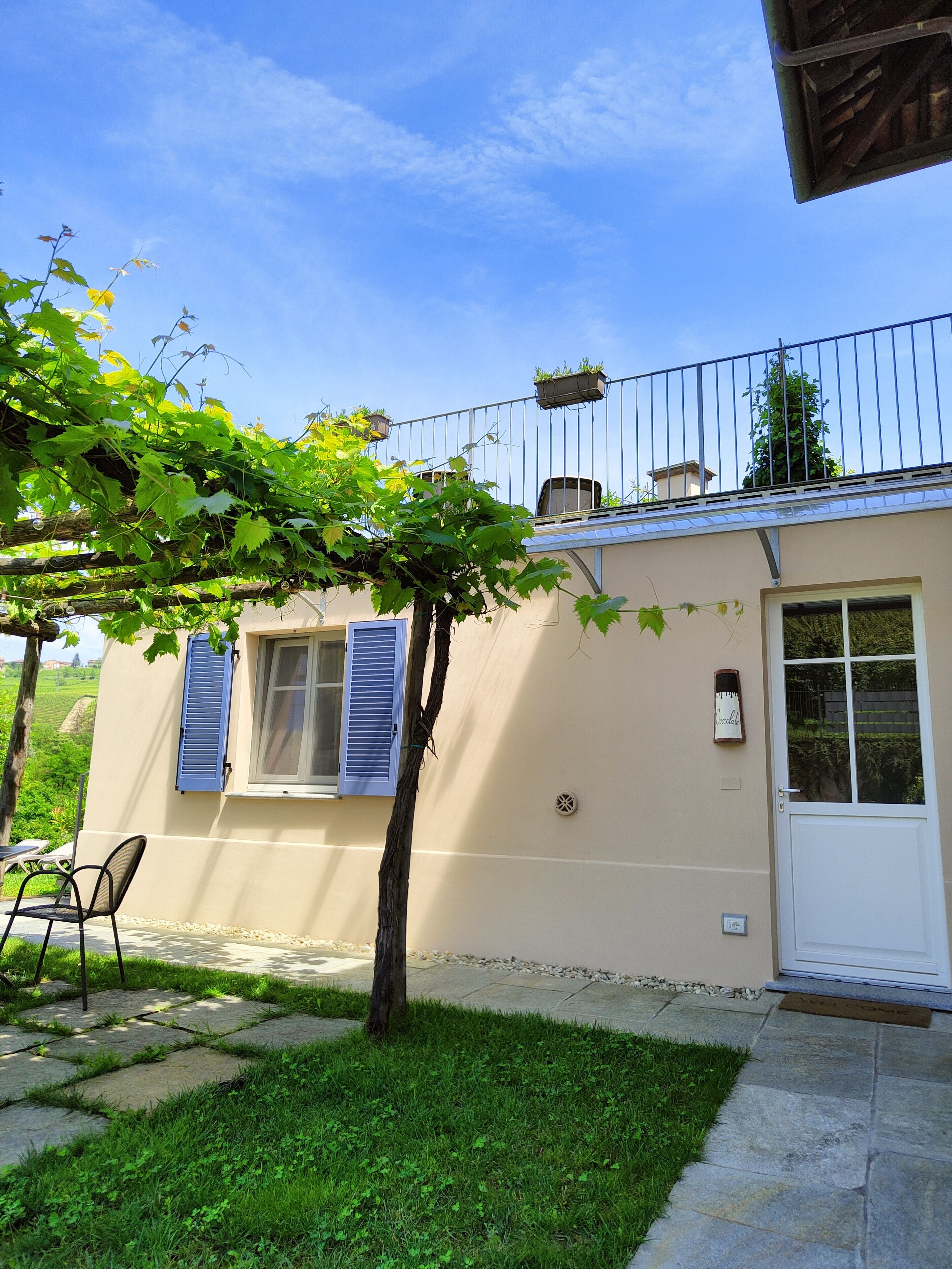 Una casa con un piccolo giardino, una pergola ricoperta di piante rampicanti, una porta bianca e una finestra con persiane blu.