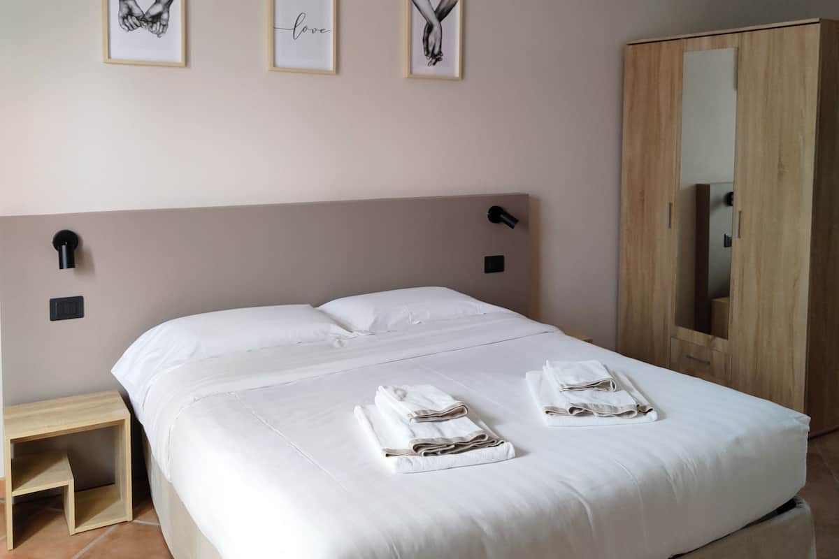 Een net opgemaakt bed met witte lakens en handdoeken, een houten nachtkastje, een wandlamp en drie ingelijste foto’s aan de muur.