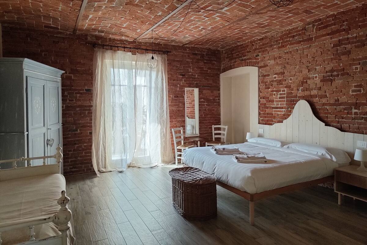 Een slaapkamer met een bakstenen muur, een houten vloer, een bed, een kledingkast en een raam met dunne gordijnen.