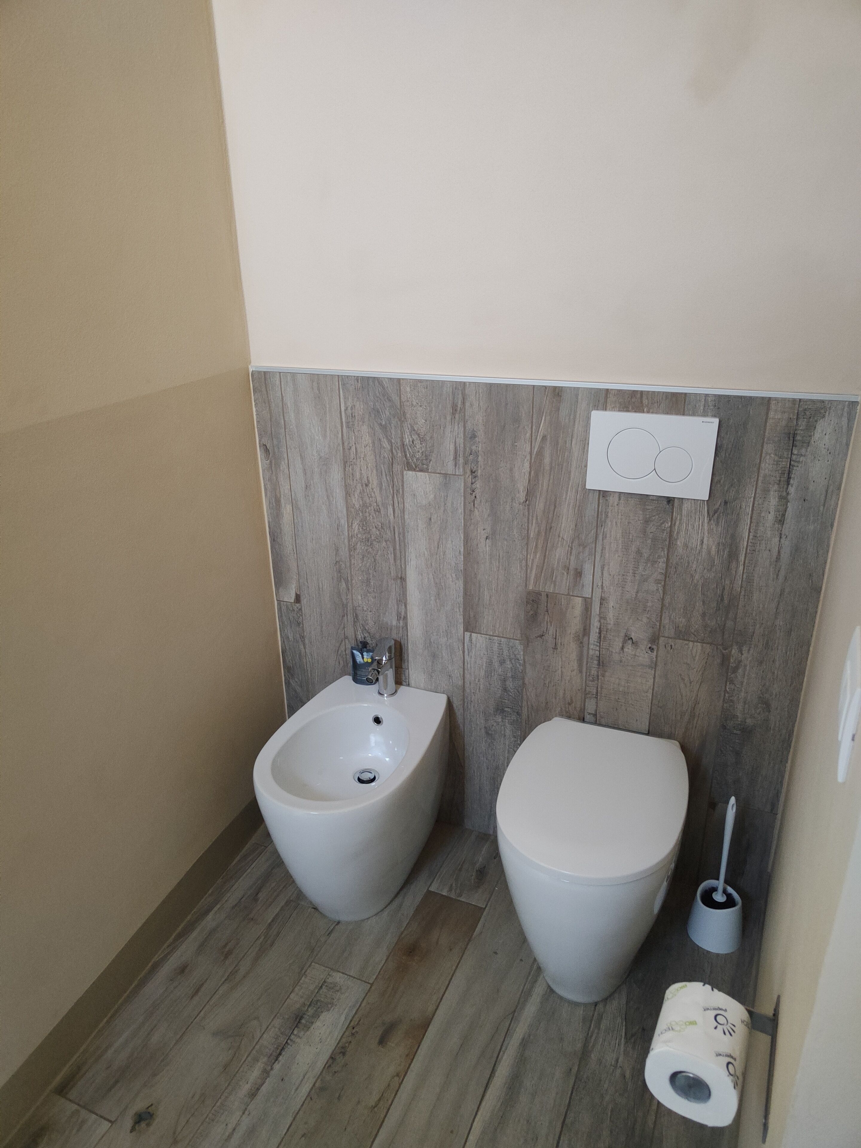A bathroom with a bidet, toilet, wooden floor, and a wall-mounted flush handle.