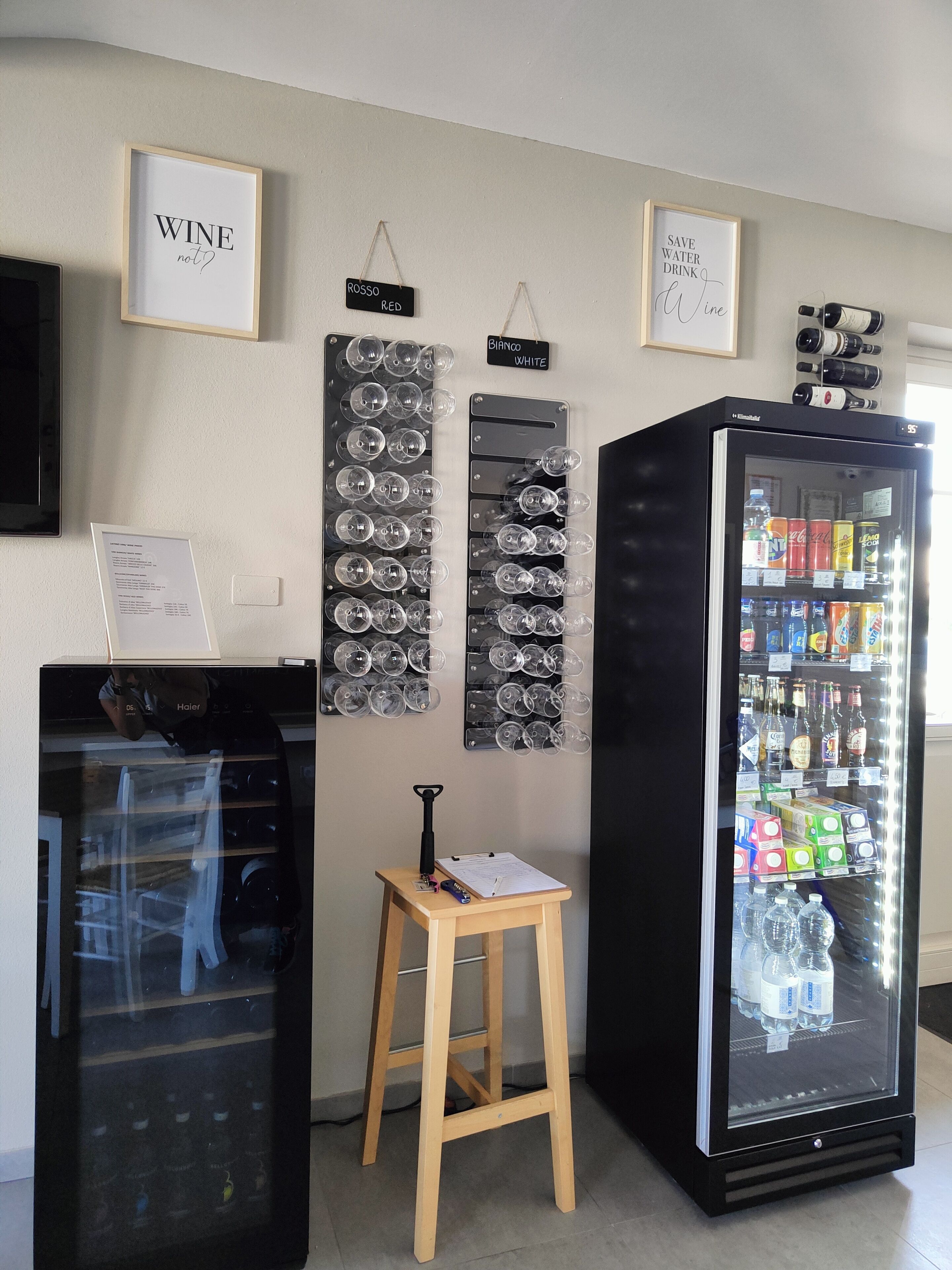 Une pièce équipée d’un réfrigérateur rempli de boissons, d’un petit tabouret sur lequel est posée une bouteille, et de verres à vin fixés au mur.