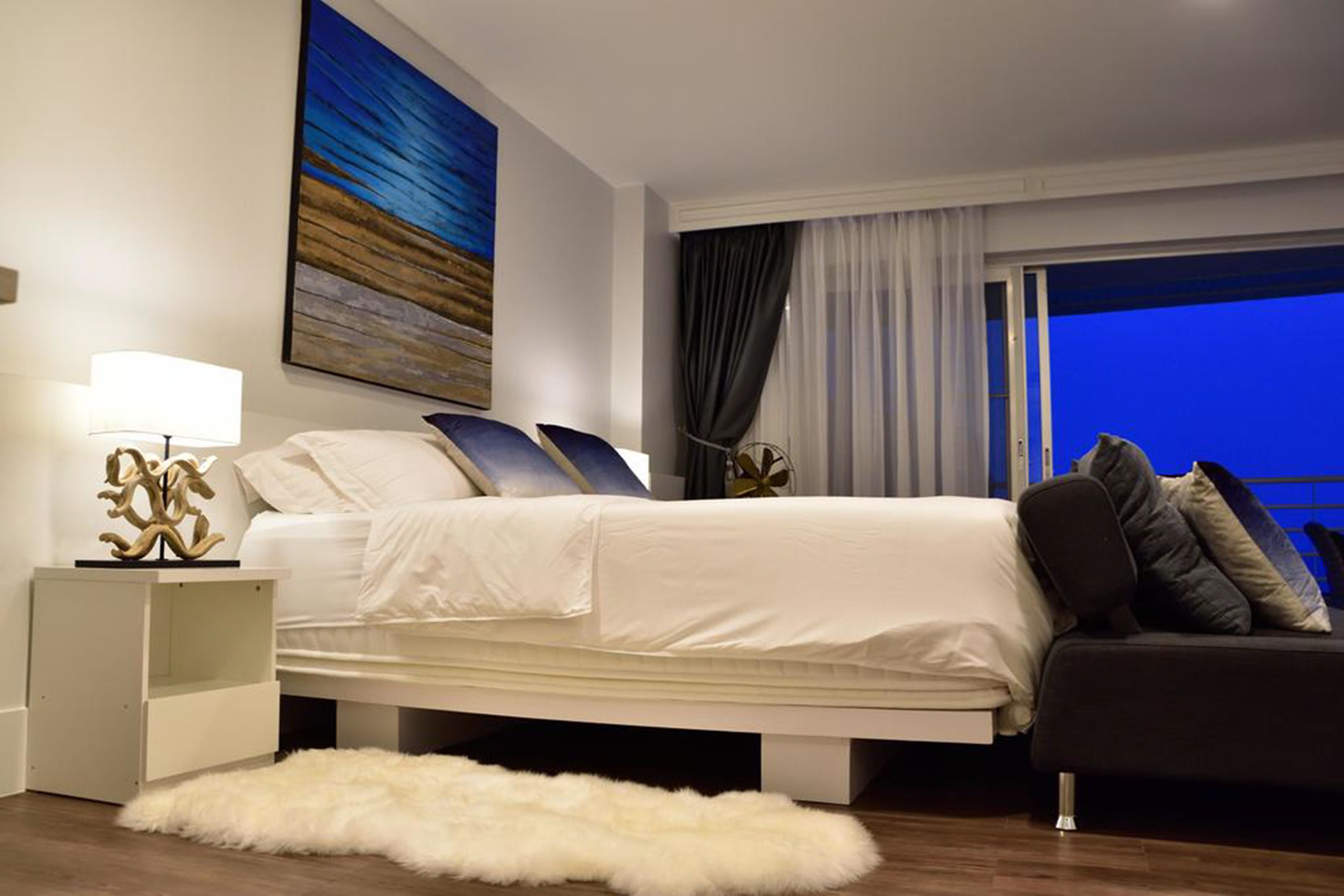 A modern hotel room with a large bed, a nightstand with a lamp, a fluffy rug, and a view of the night sky through a window.