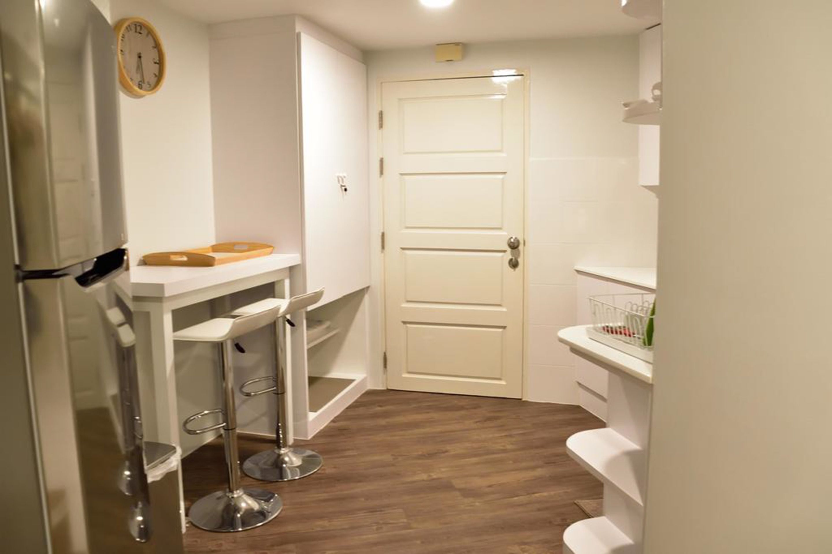 A compact kitchen with a white door, a refrigerator, a small bar stool, and a shelf with kitchen items.