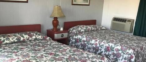 Habitación de hotel con dos camas, una mesita de noche, una lámpara, un calentador y dos cuadros en la pared.