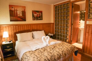 Habitación de hotel con cabecera de madera, cama con colcha estampada, dos cuadros en la pared y una puerta de madera con panel de cristal.