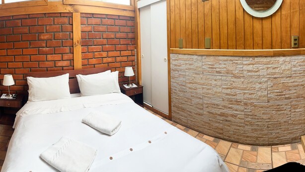 Quarto de hotel com parede de tijolos, parede de pedra e teto de madeira. Há duas camas com roupa de cama e toalhas brancas.