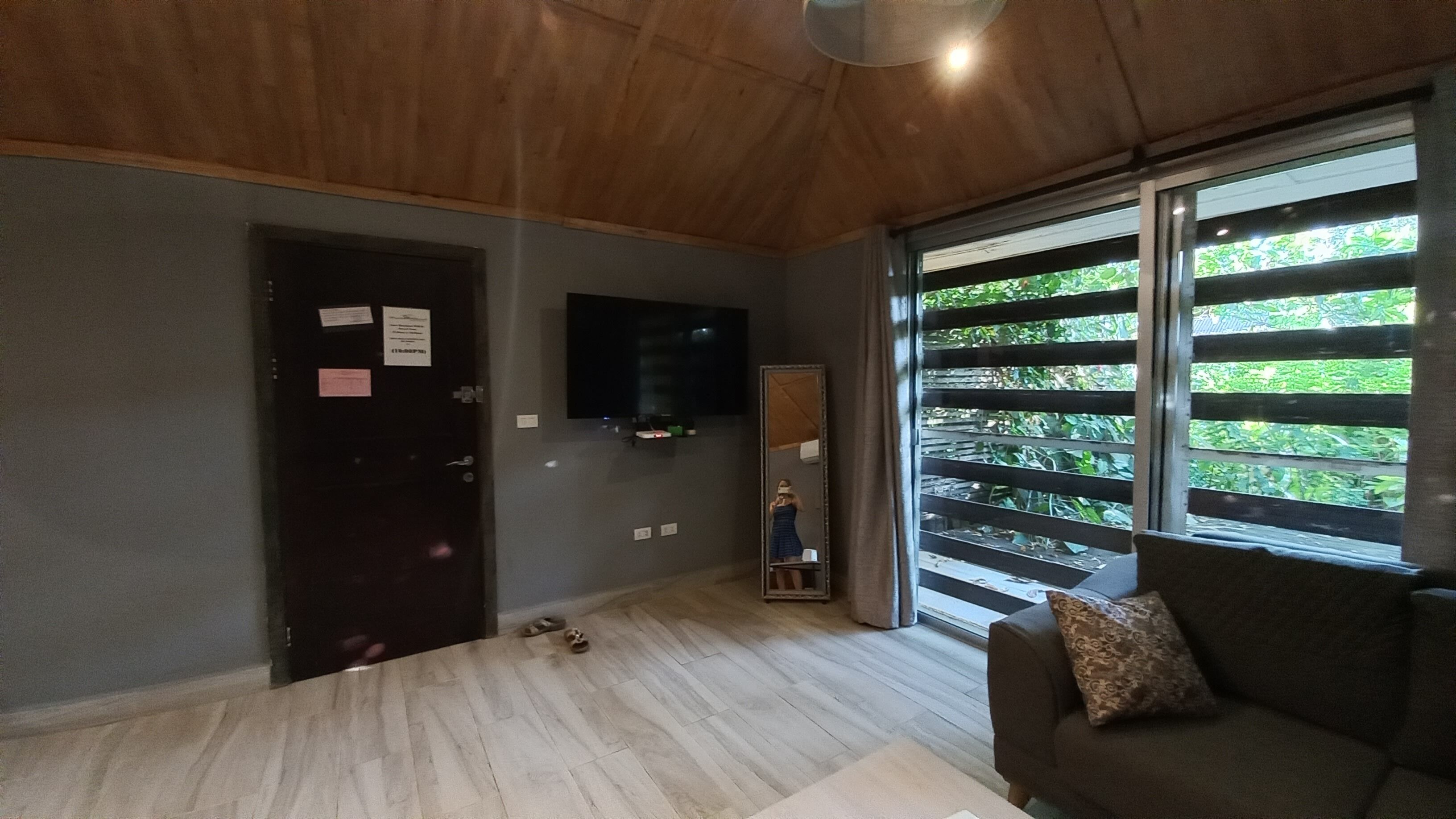 Habitació amb sostre de fusta, porta fosca plena de papers, televisió de pantalla plana, finestra gran amb persianes, sofà i mirall.