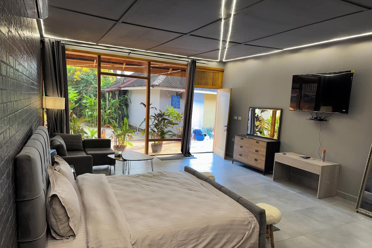 A modern bedroom with a large bed, a desk, a TV, and a balcony with plants.