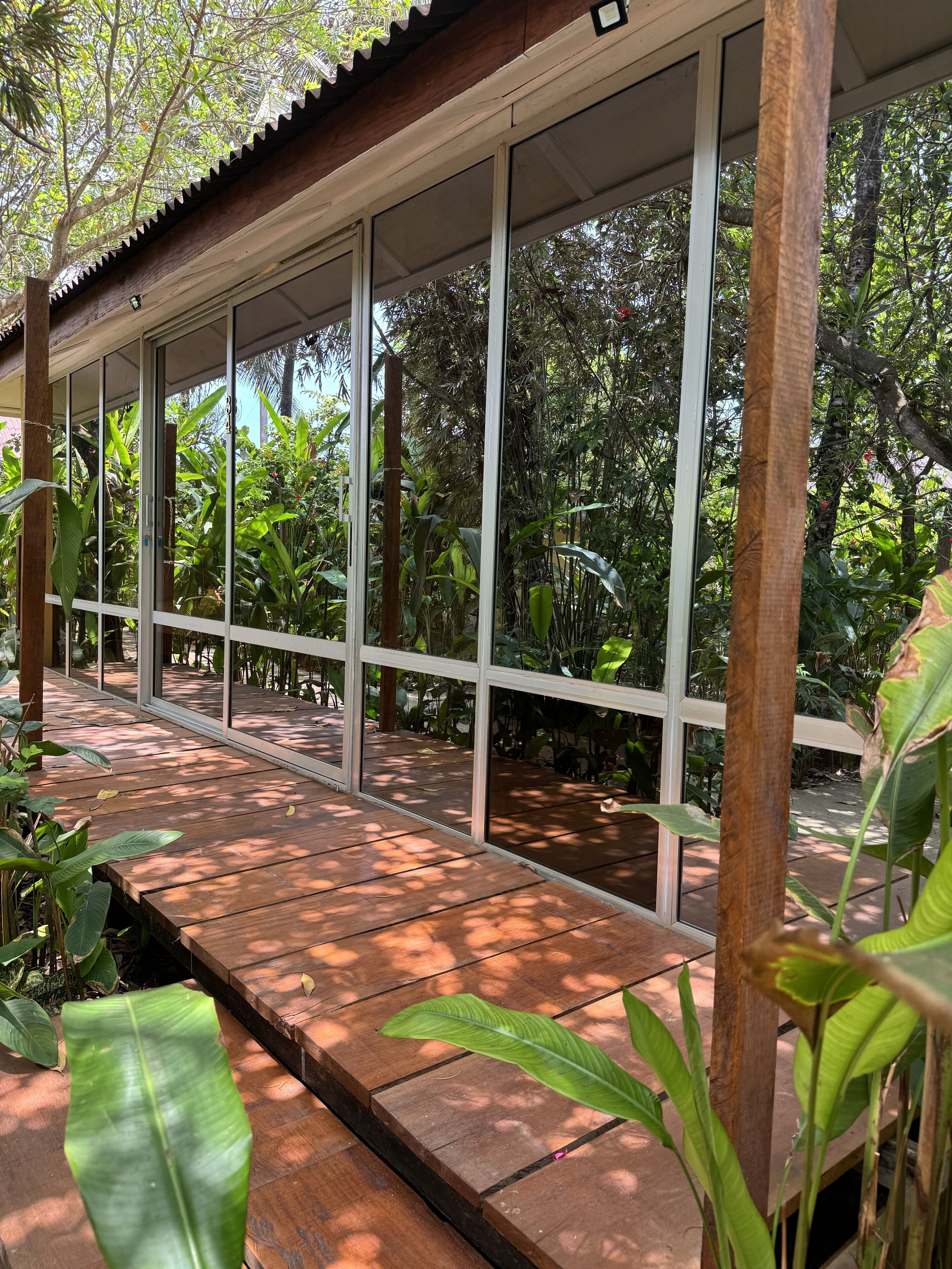 Una terraza cubierta de madera con amplios ventanales de vidrio, rodeada de abundante follaje.