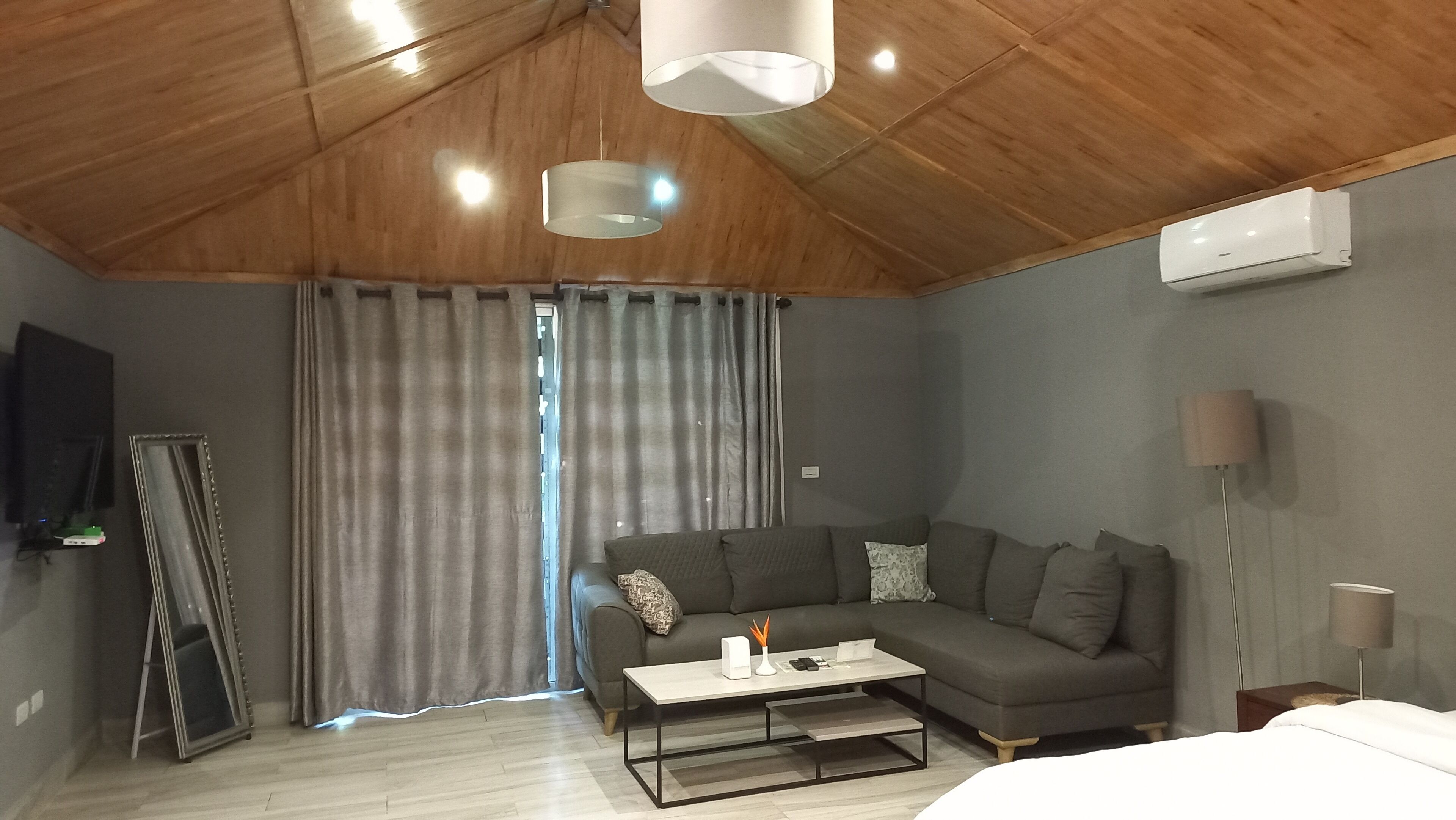 A modern living room with a wooden ceiling, grey curtains, a grey sofa, a white coffee table, and a flat-screen TV mounted on the wall.