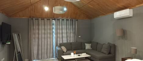 A modern living room with a wooden ceiling, grey curtains, a grey sofa, a white coffee table, and a flat-screen TV mounted on the wall.