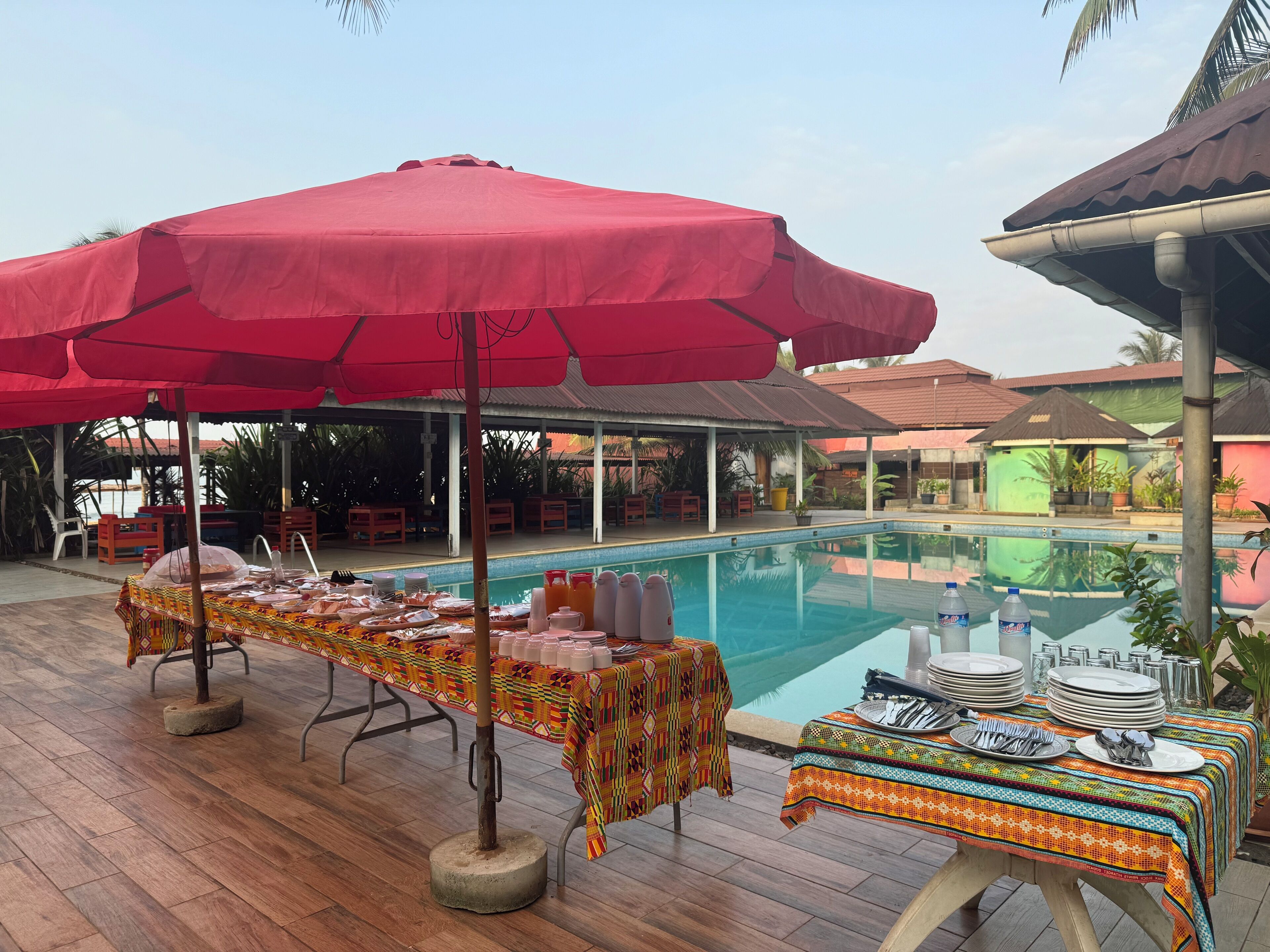 Zona junto a la piscina con mesas dispuestas para comer, cubierta por sombrillas rojas, y una piscina al fondo.