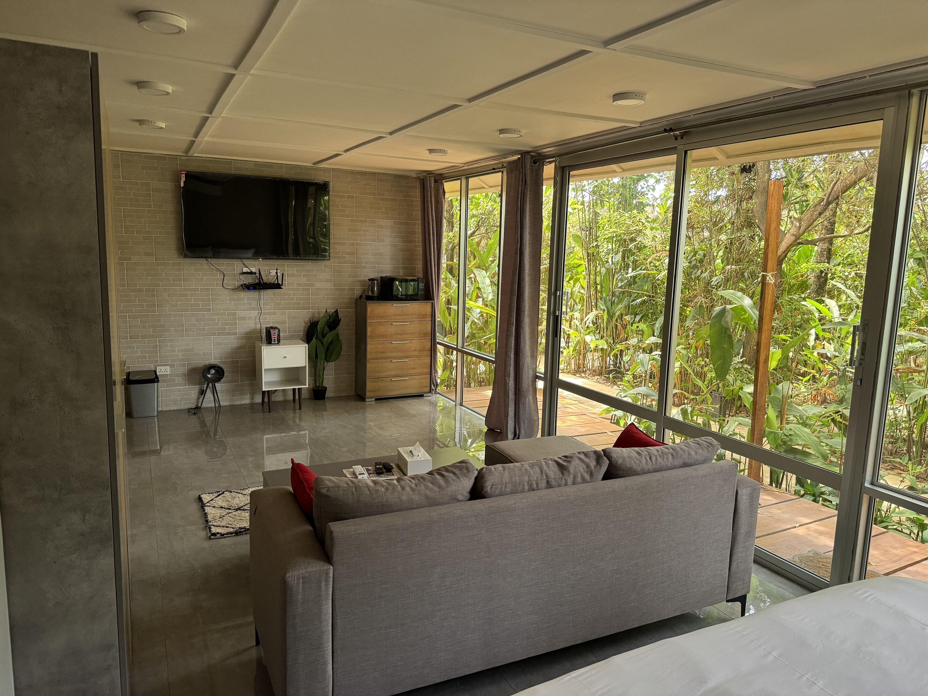 A modern living room with a grey sofa, a flat-screen TV mounted on the wall, a wooden cabinet, and a large window offering a view of greenery.
