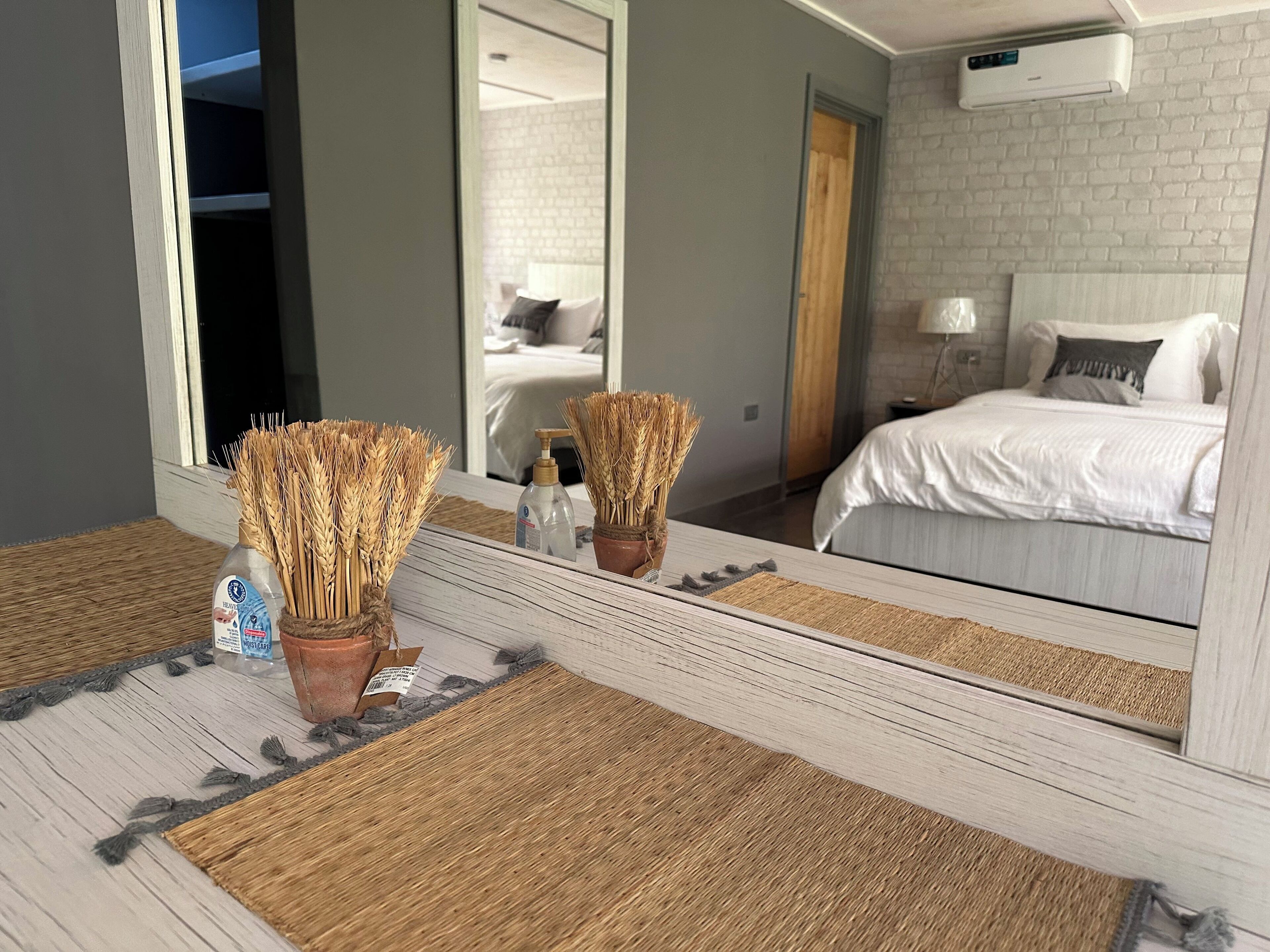 A hotel room with a large mirror reflecting a bed, a bedside table with a bottle and a vase of dried plants, and a wooden floor.