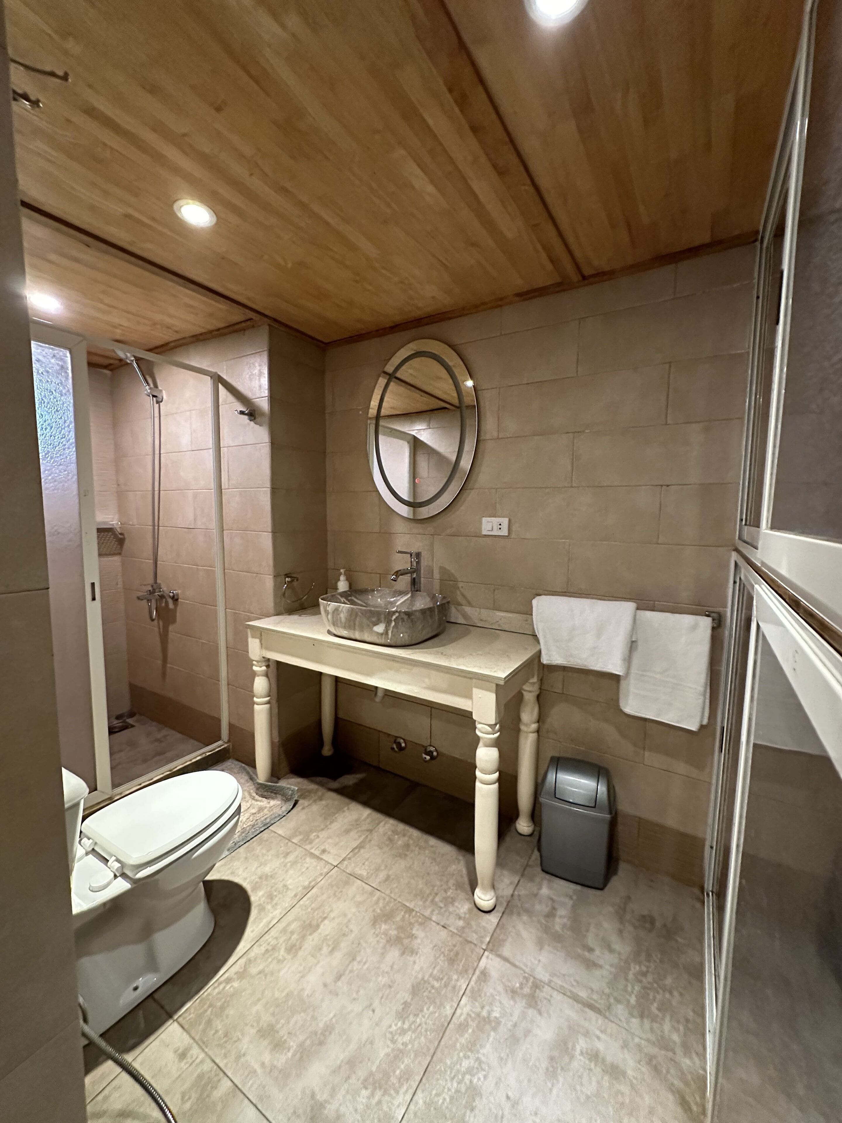 A bathroom with a wooden ceiling, a round mirror, a sink, a toilet, and a shower.