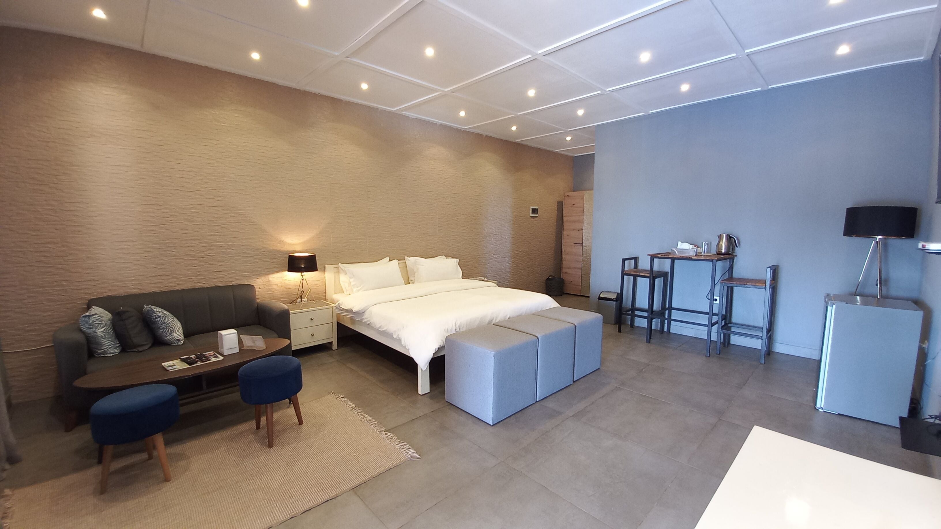 A modern hotel room with a bed, a desk, a sofa, and a small kitchenette.