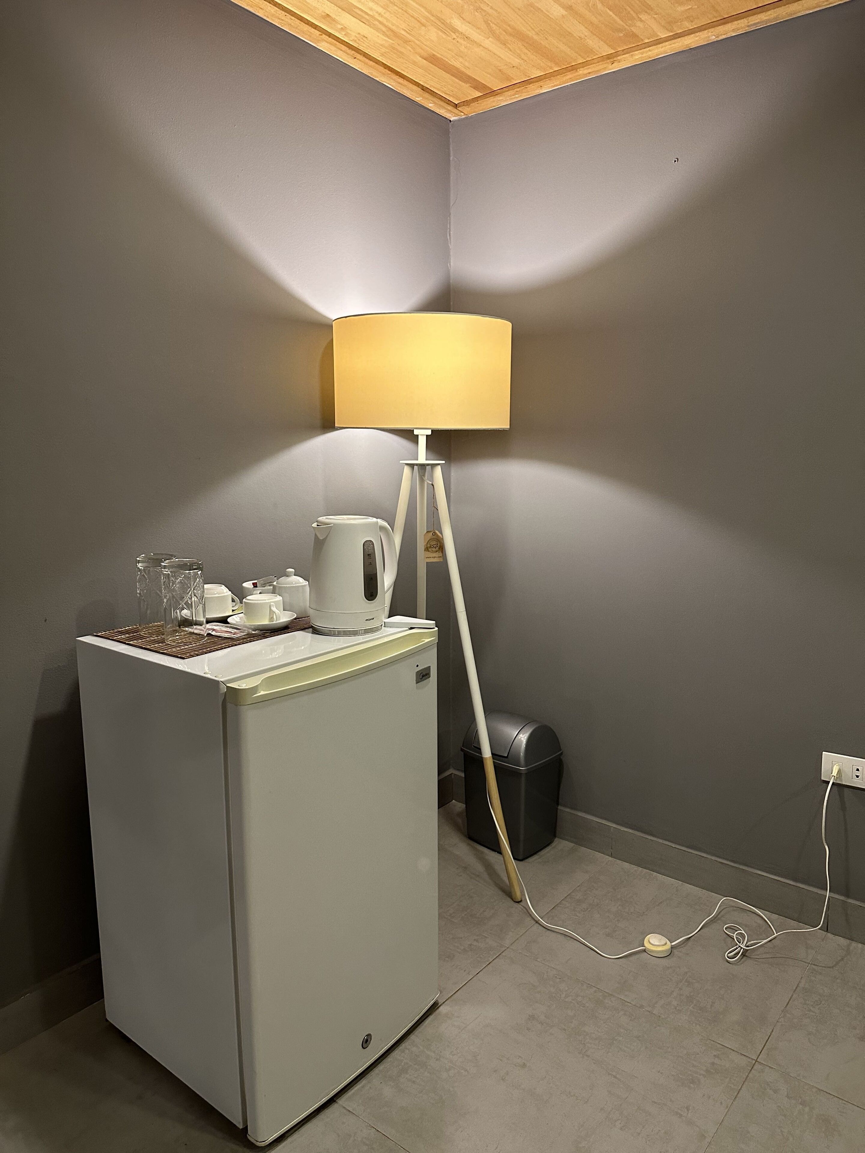 Ett litet, praktiskt kök med ett kylskåp, en kaffebryggare och en vattenkokare på bänkskivan. En golvlampa med en vit lampskärm står placerad intill väggen.