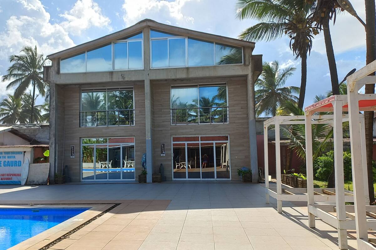 A two-story building with large windows, a swimming pool, and palm trees in the background.