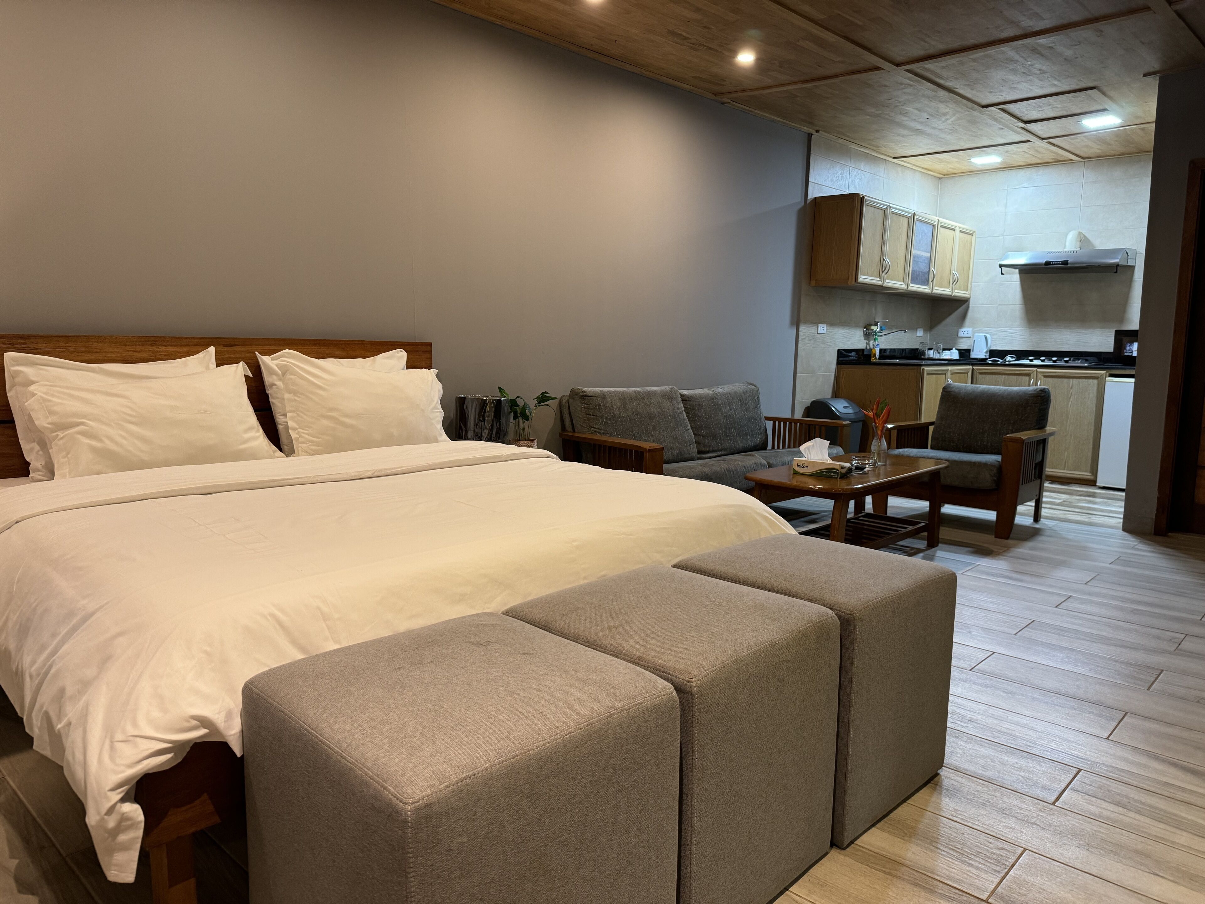 A hotel room with a large bed, two armchairs, a small table, and a kitchenette in the background.