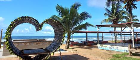 A beachside area with a heart-shaped structure, palm trees, and a covered seating area with a view of the ocean.