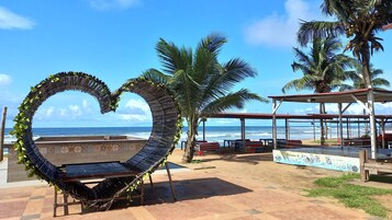 Zona de playa con una estructura en forma de corazón, palmeras y un área de estar cubierta con vista al océano.