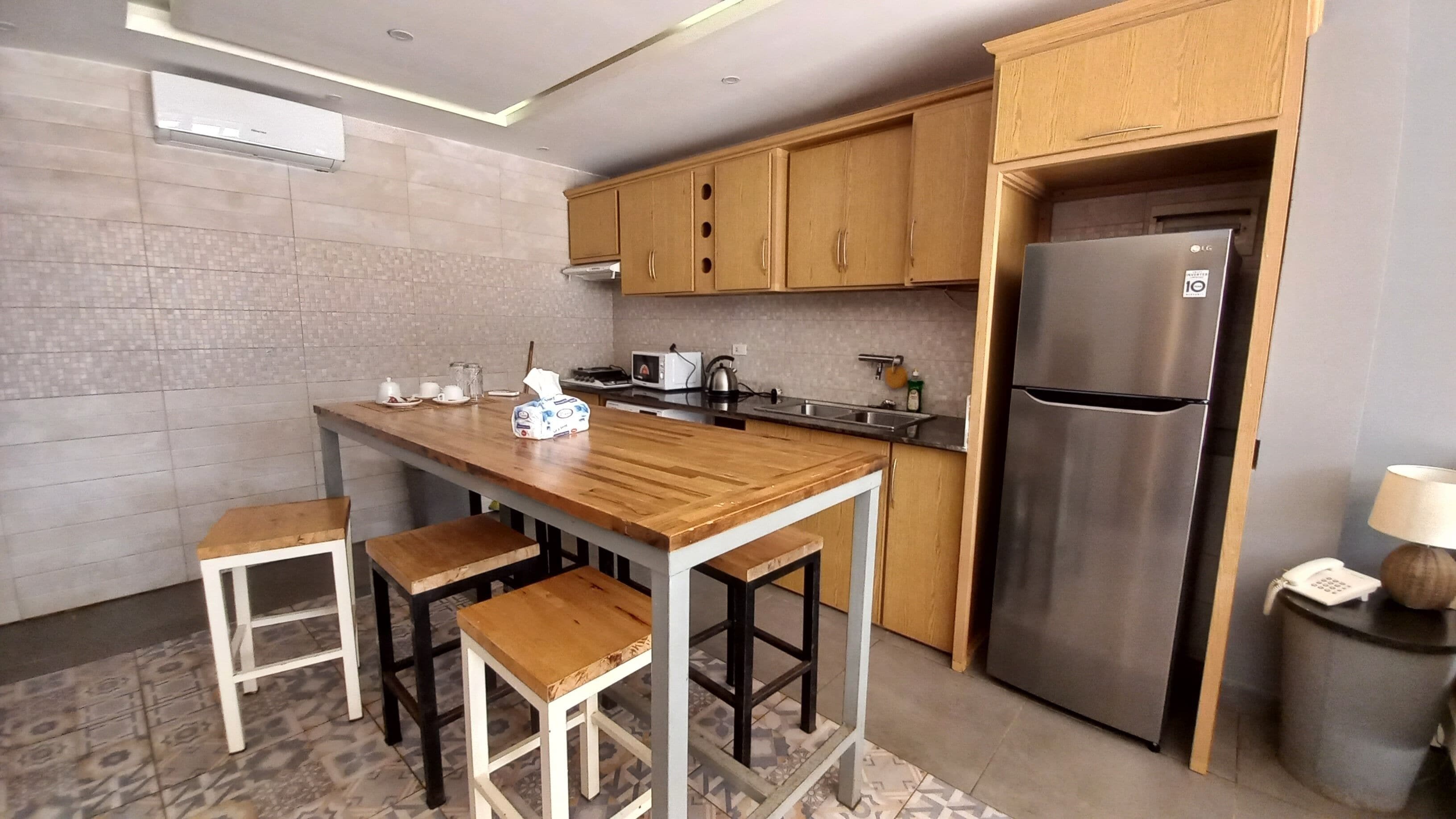 A modern kitchen with wooden cabinets, a stainless steel refrigerator, a wooden dining table with bar stools, and a tiled backsplash.