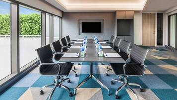 A conference room with a long table, multiple chairs, water bottles, a flat-screen TV, and large windows with a view of greenery.