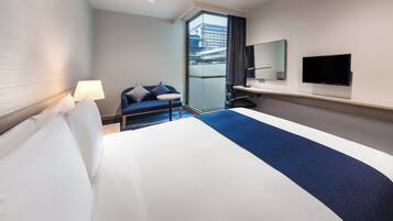 Une chambre d’hôtel avec un lit, un bureau, une chaise, un téléviseur et une vue sur un immeuble, visible à travers une porte vitrée.